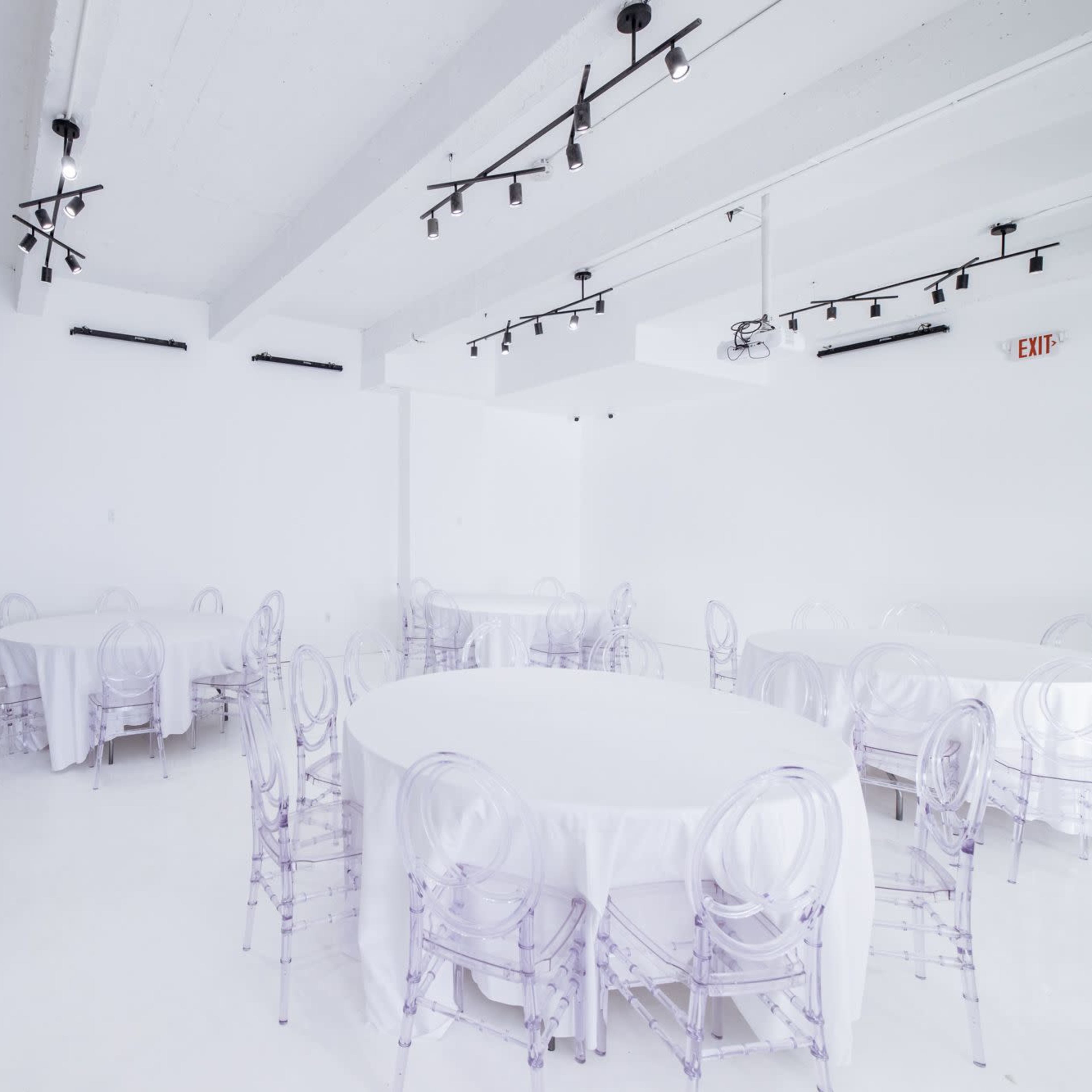 A spacious, white event room with several circular tables covered in white tablecloths and surrounded by clear plastic chairs.