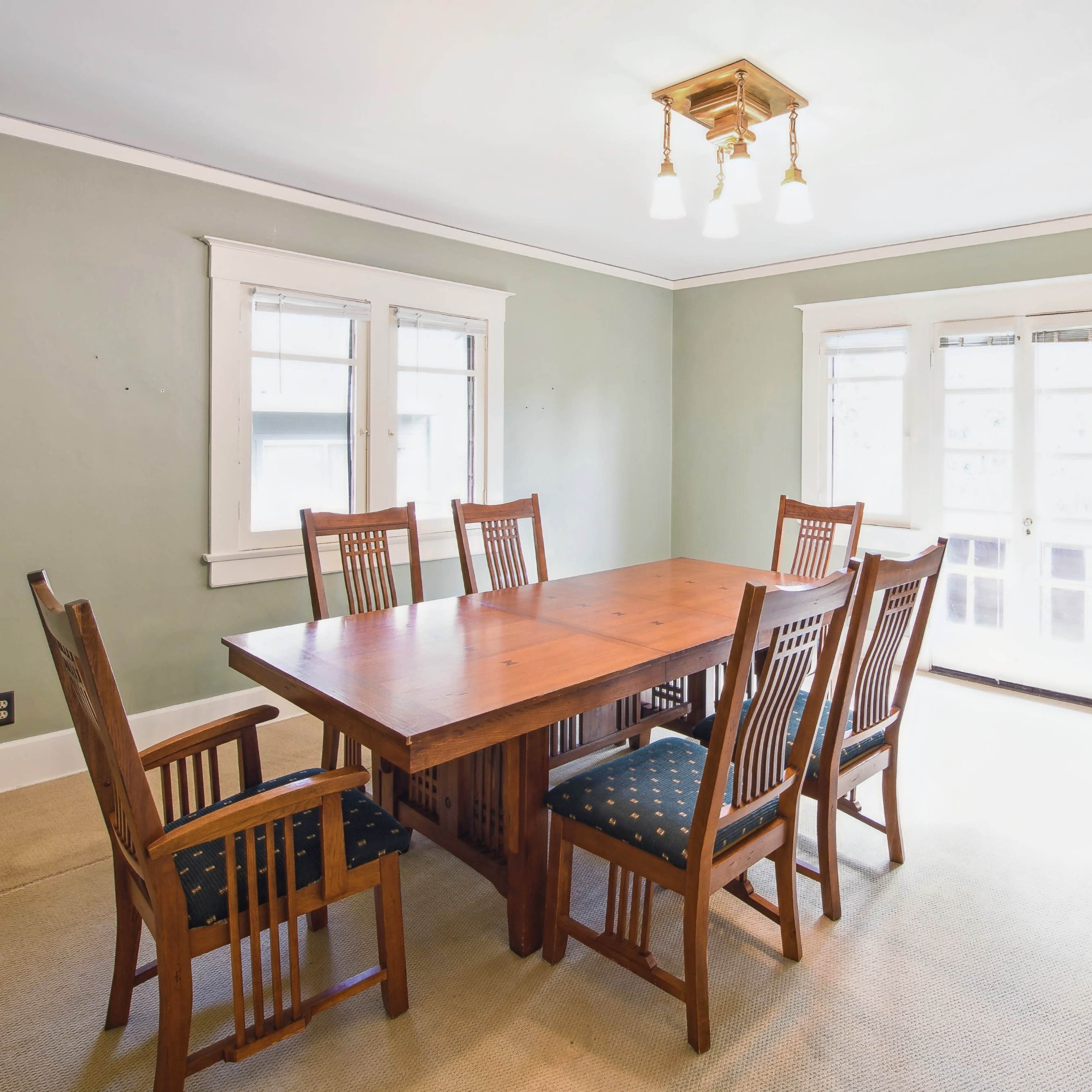 A dining room features a wooden table surrounded by six chairs, with windows illuminating the space and a sideboard positioned against the wall.
