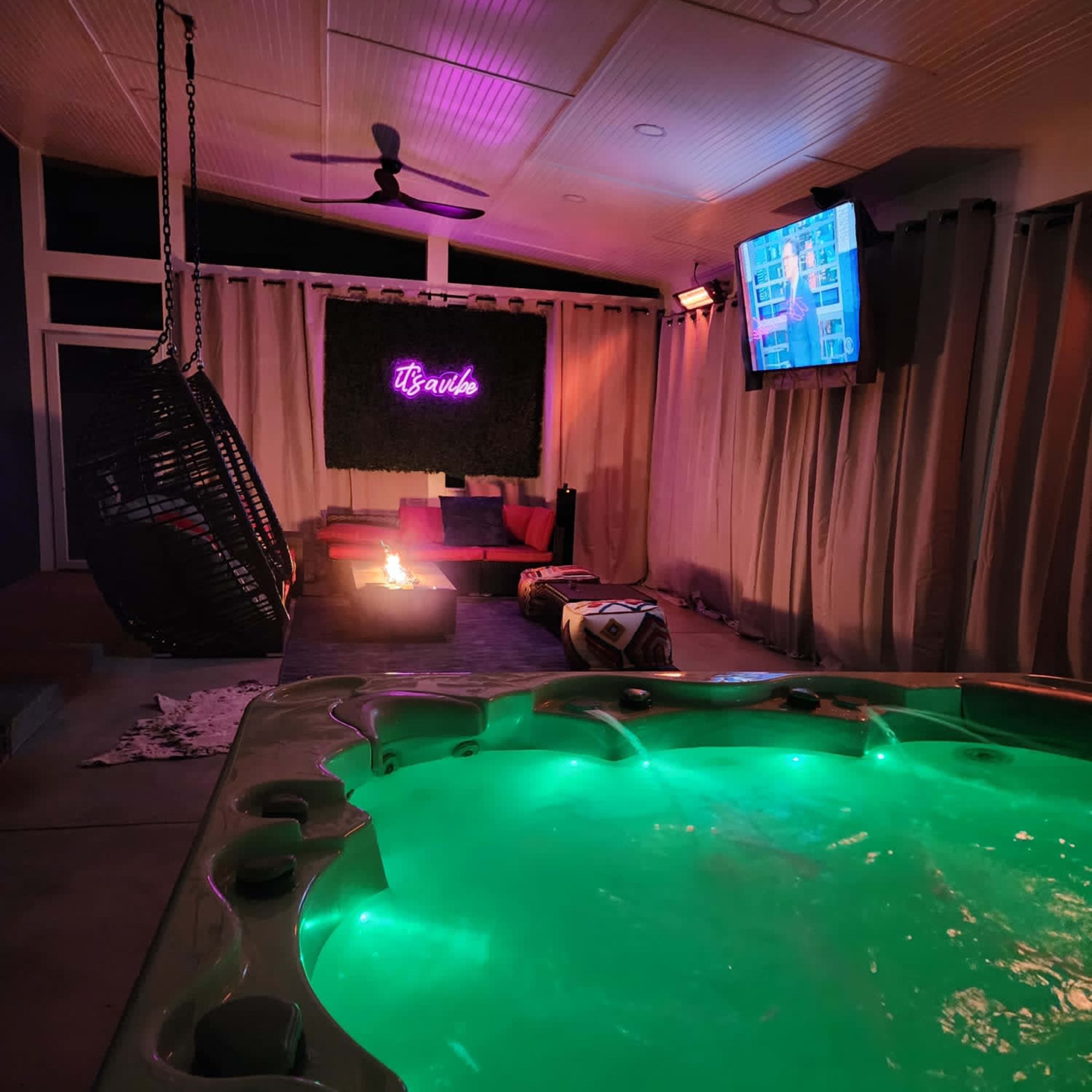 A lit hot tub with green water is set in a cozy indoor space featuring a hanging chair, a television, and a fire pit surrounded by dim lighting.
