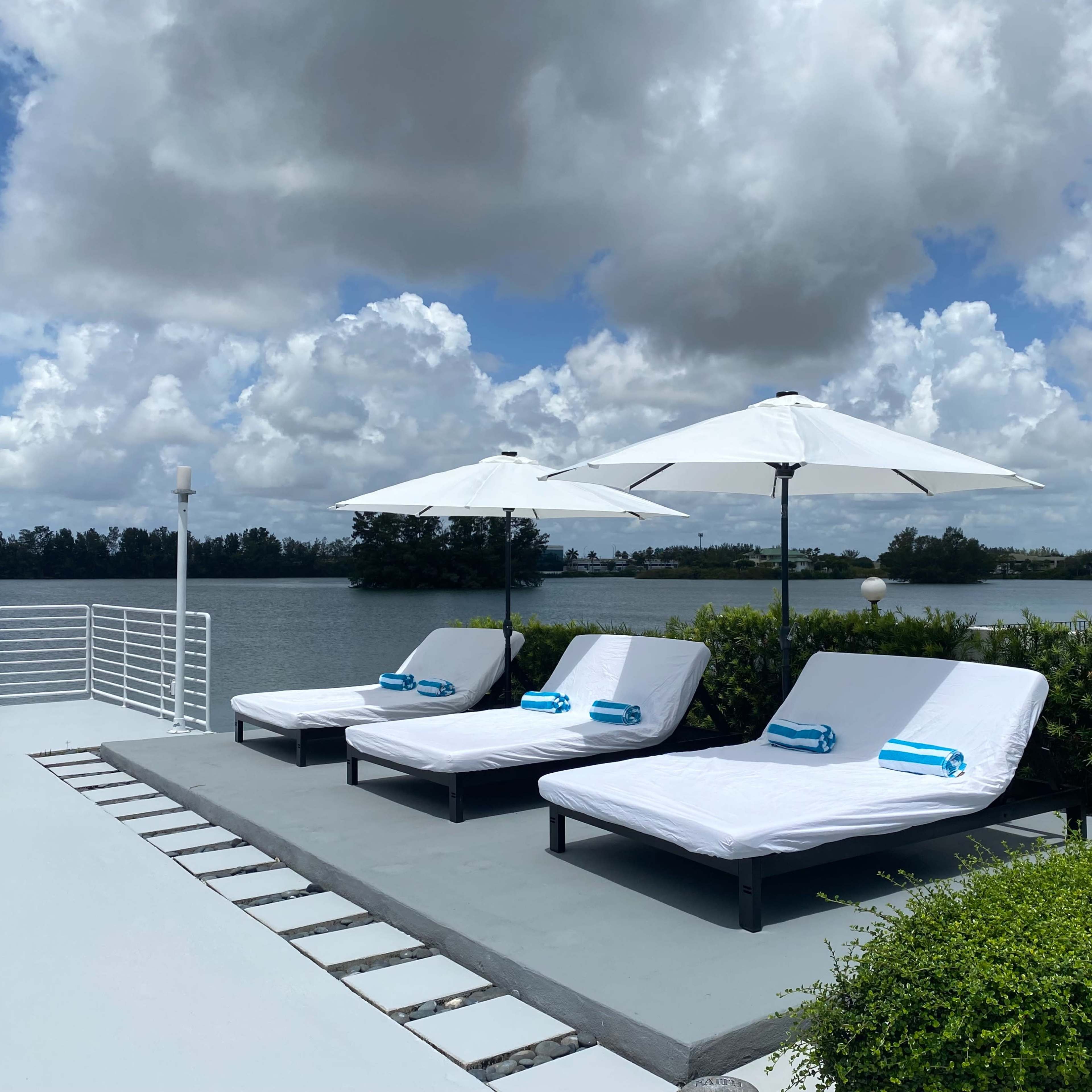 Three lounge chairs with blue towels sit under white umbrellas by a river with greenery in the background.