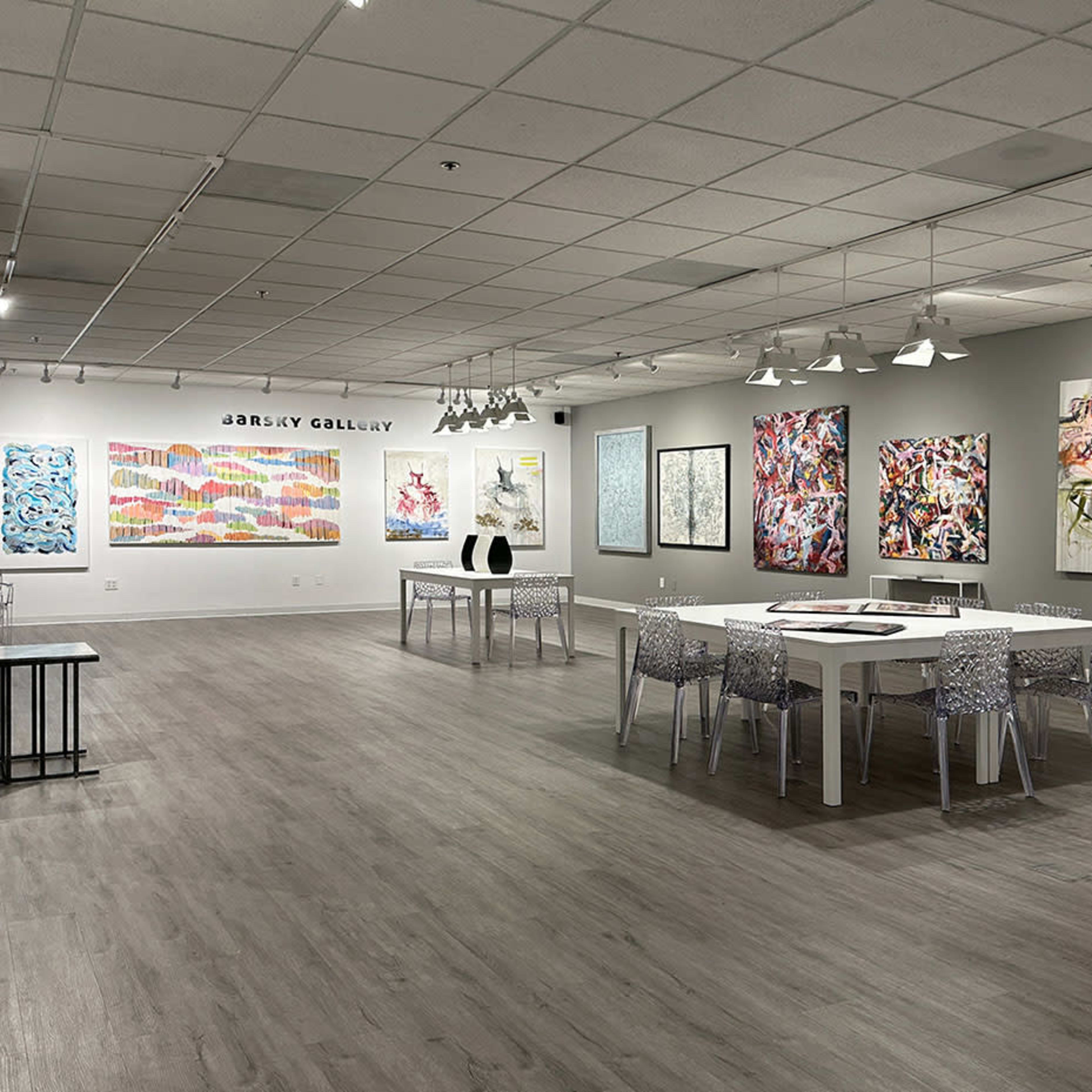 A spacious art gallery contains various contemporary paintings displayed on the walls and a few tables with chairs in the center.