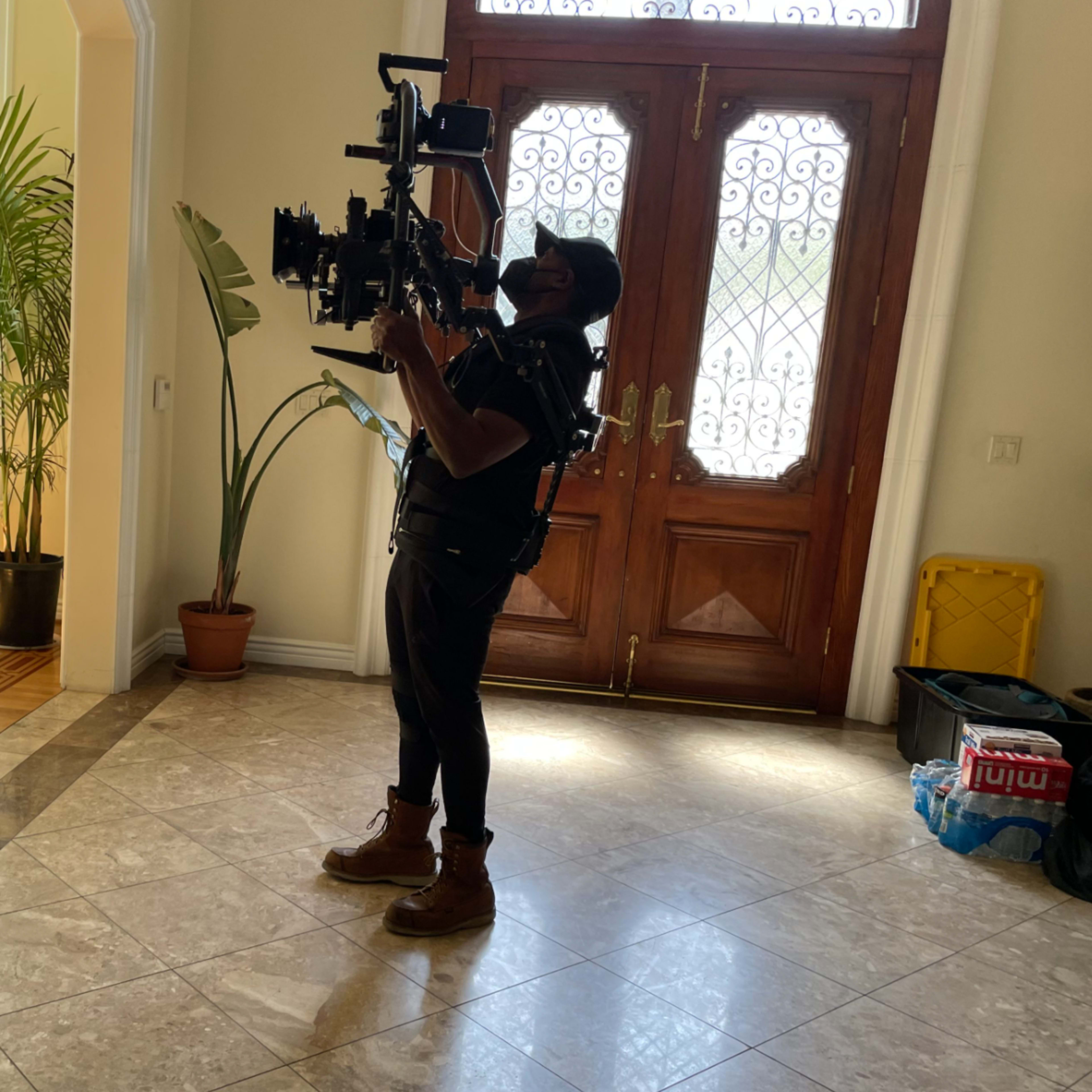 A person holding a camera rig stands inside a room with large front doors and a plant in a pot.