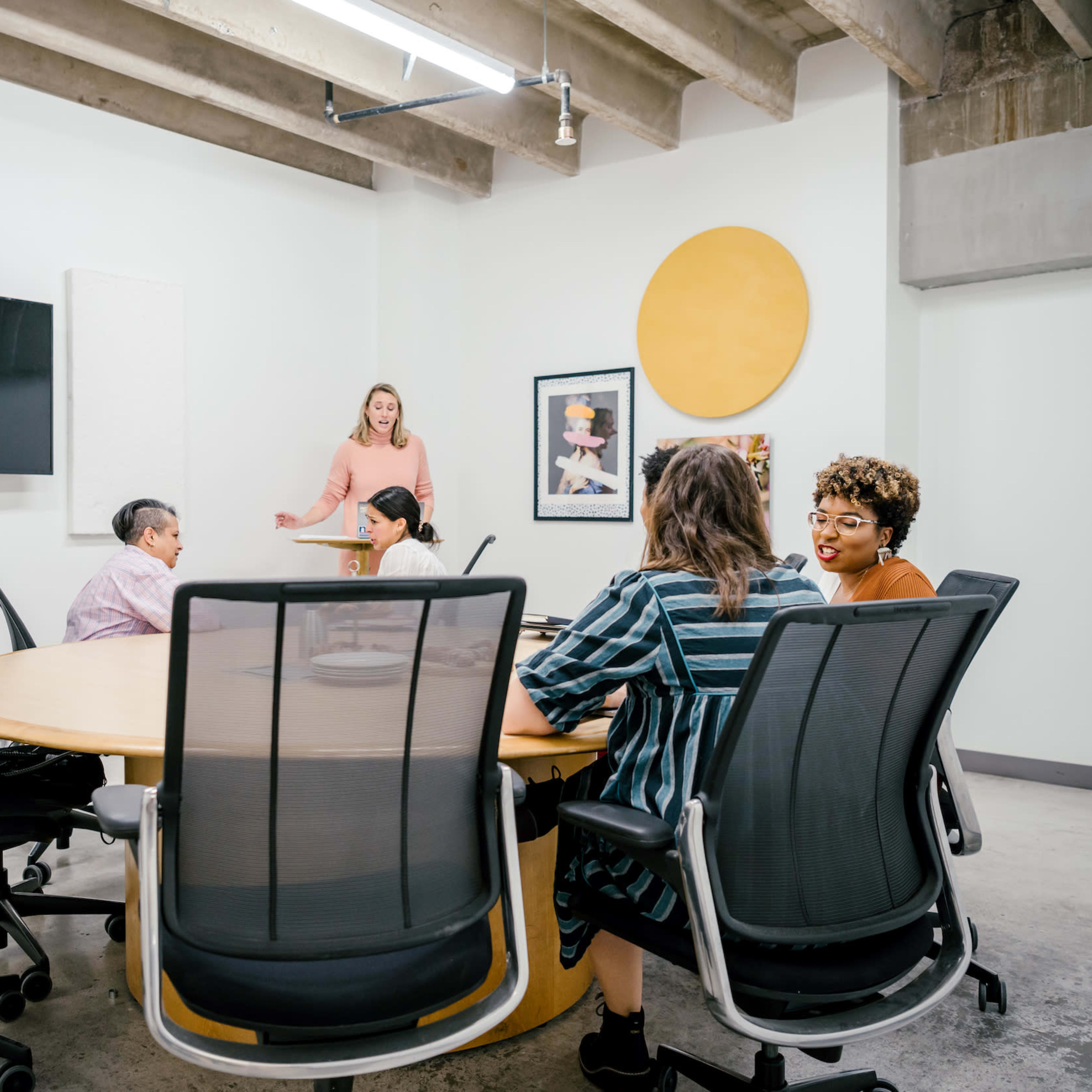 A woman stands at the front of a conference room with a round table surrounded by several people engaged in discussion.