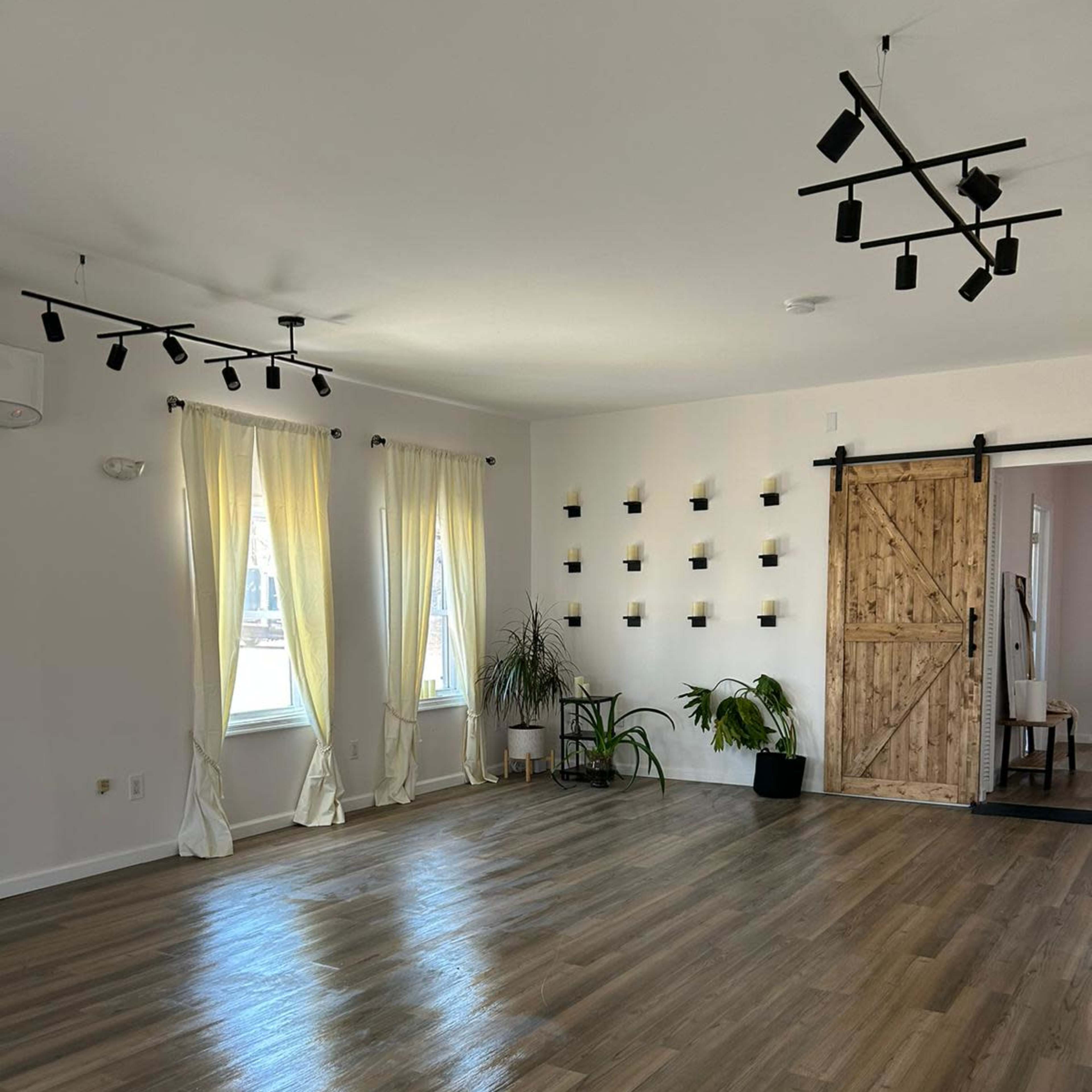 A spacious, well-lit room with wooden flooring, large windows draped with light curtains, and a wooden sliding door.