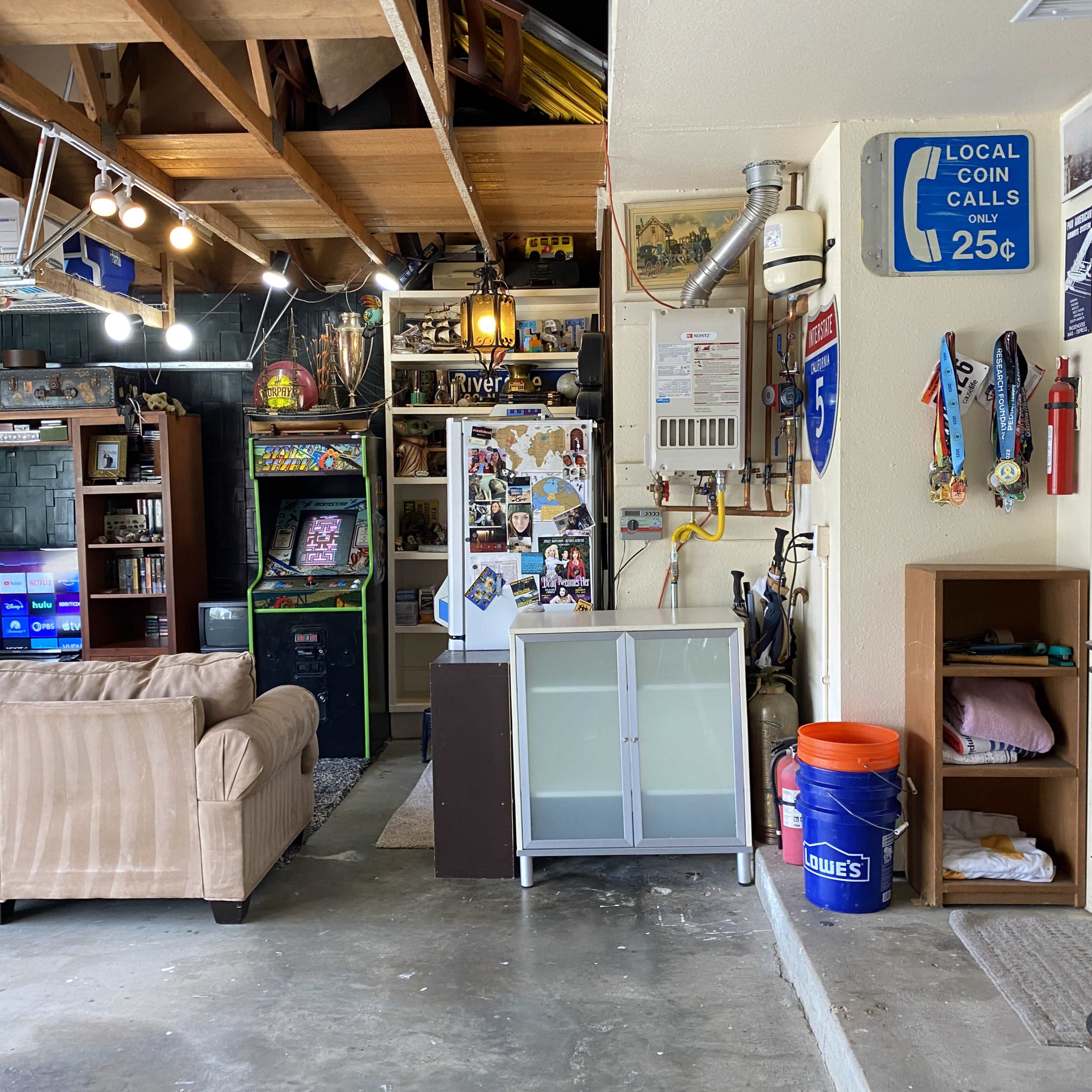 A garage space featuring a couch, a refrigerator, an arcade machine, and various decorations, with a sign indicating local coin calls.