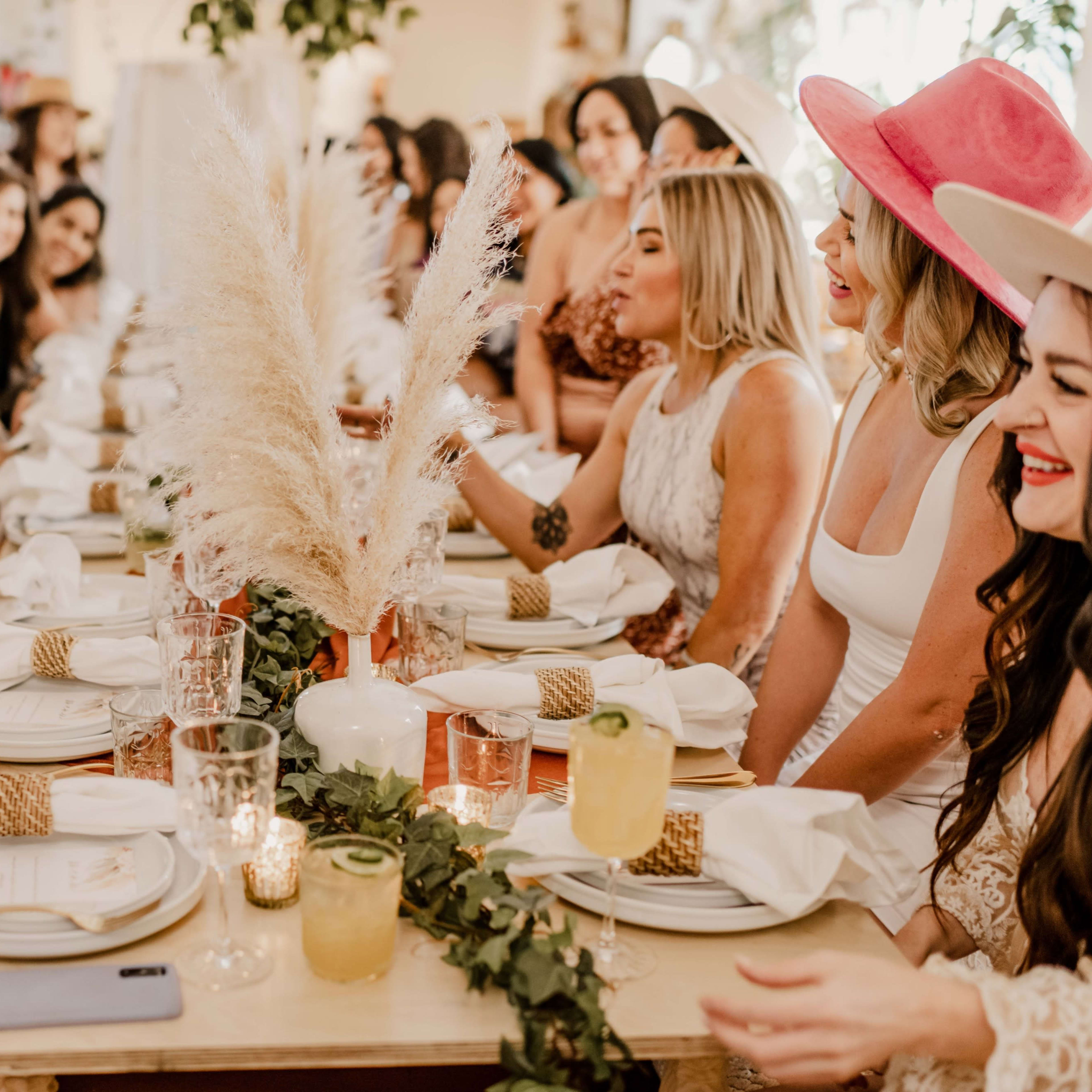 A large group of women wearing various outfits is seated around a long dinner table adorned with greenery and decorative items, engaged in lively conversation.