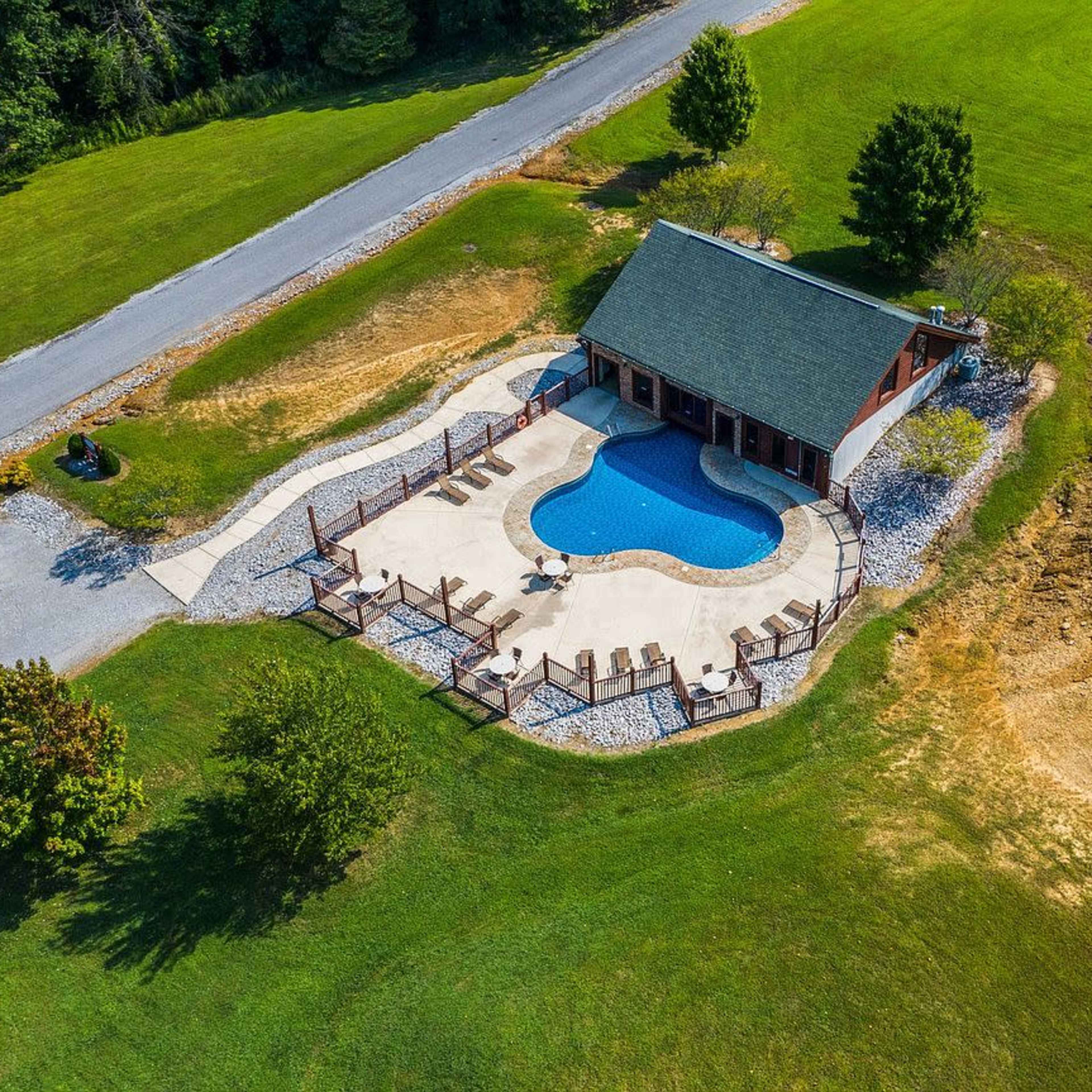 An aerial view of a house with a swimming pool surrounded by a patio and loungers, set on a green lawn near a road.