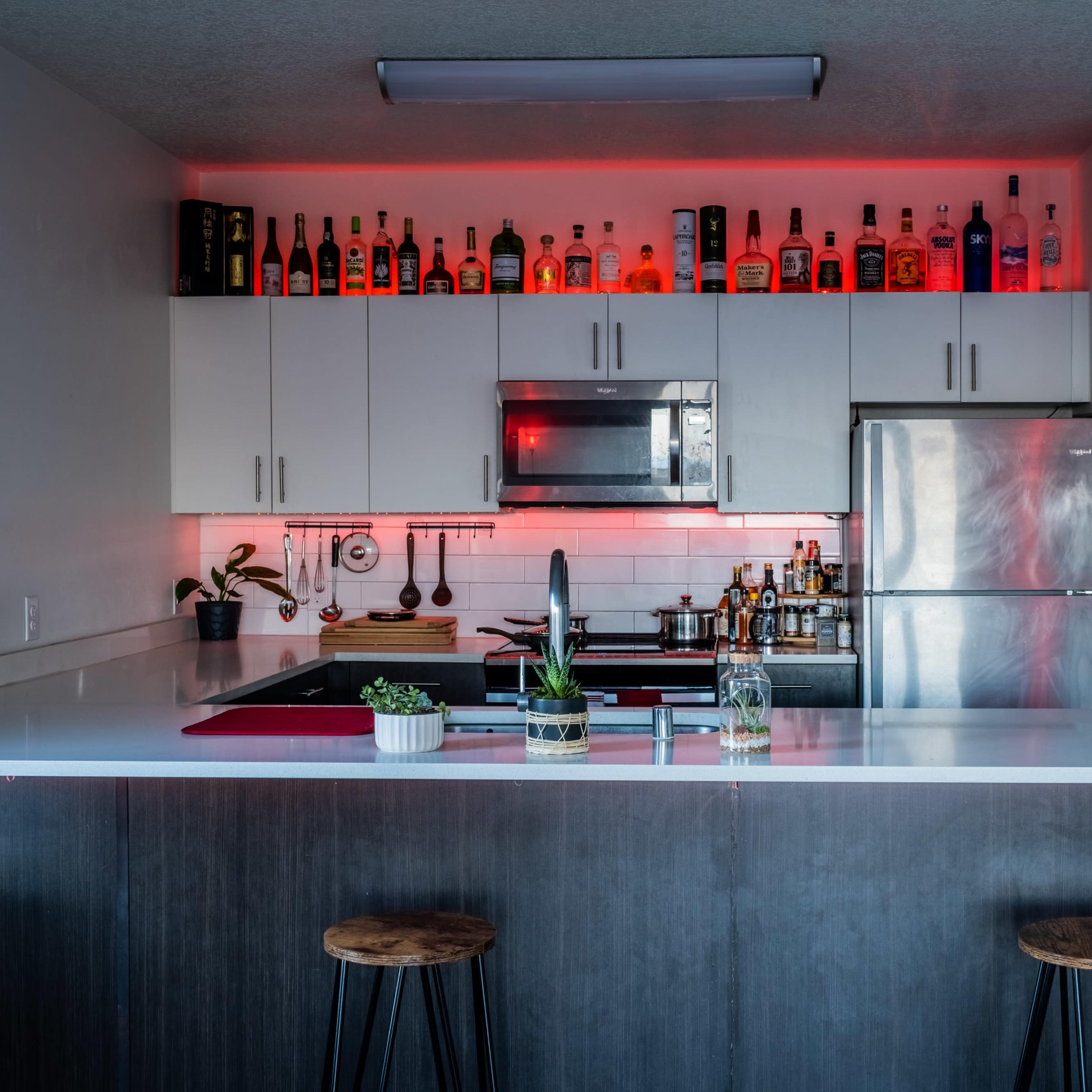The kitchen features a modern design with a white countertop, stainless steel appliances, and a row of bottles illuminated by red lighting along the upper cabinets.