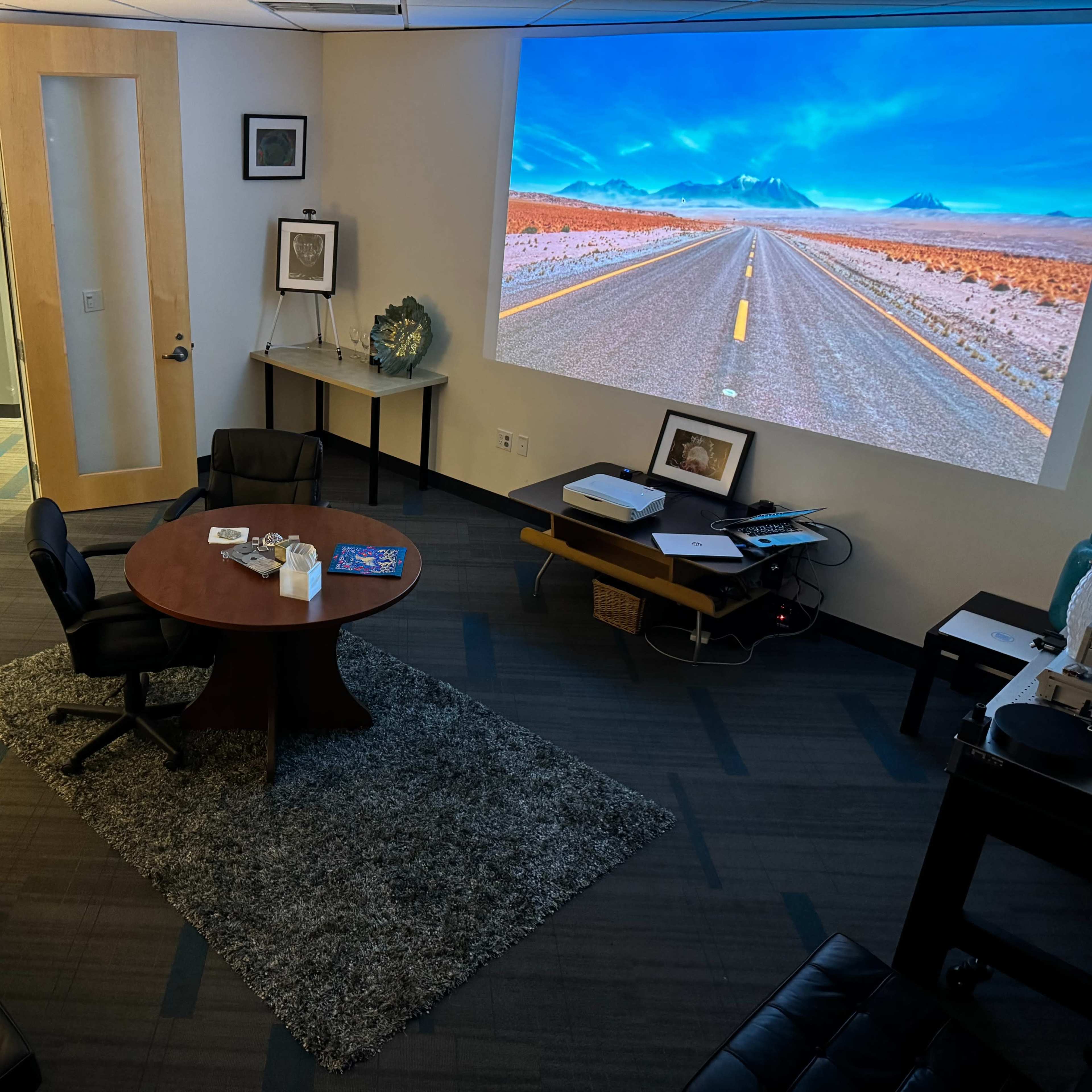 A modern office space features a round table with chairs, a large projection of a road scene on the wall, and minimalist furnishings.