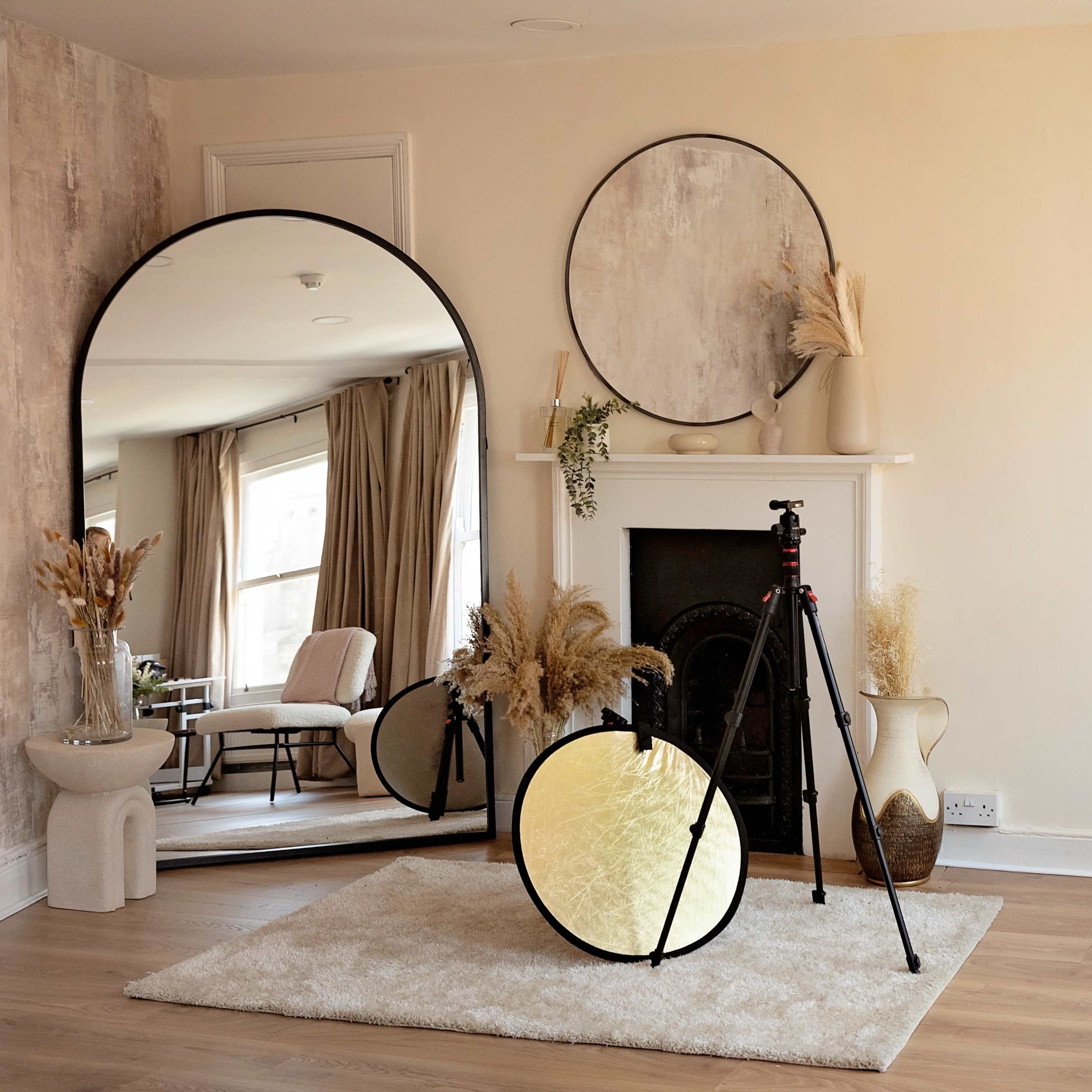 A spacious room features a large round mirror, a tripod with a reflector, and decorative elements arranged around a fireplace.