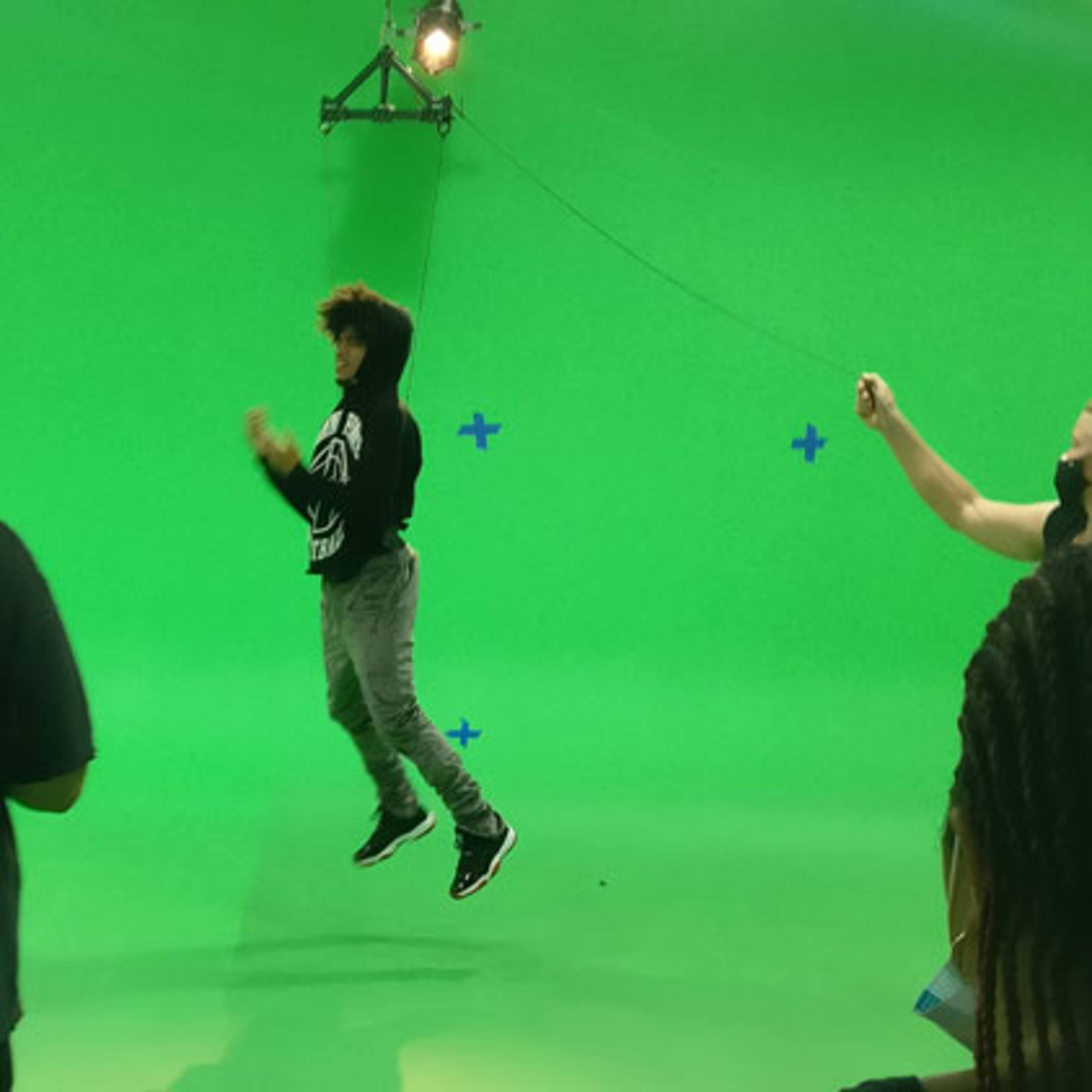 A person jumps in front of a green screen while a crew operates equipment and observes the scene.