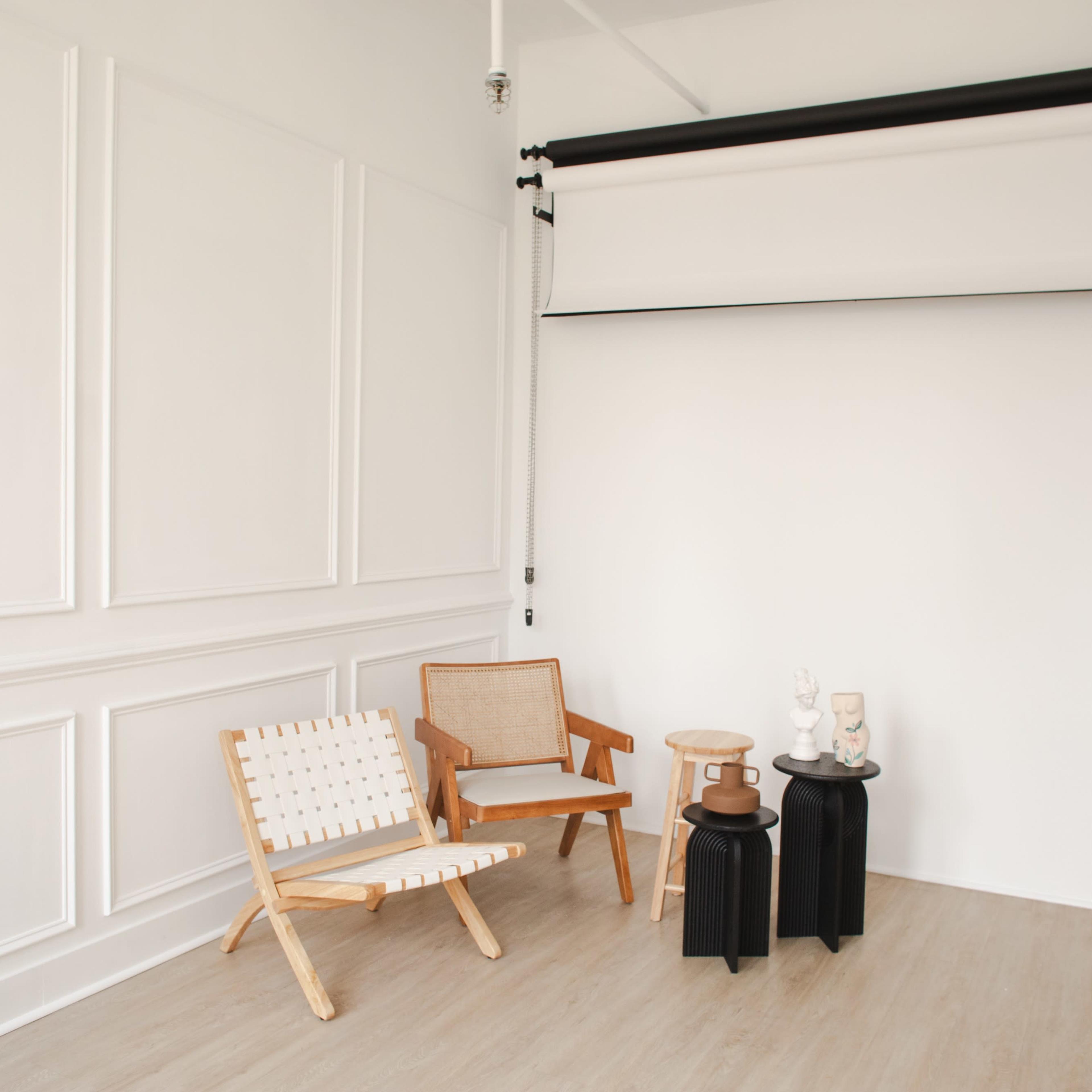 A minimalist interior room with two wooden chairs, a stool, and two small tables against a white wall.