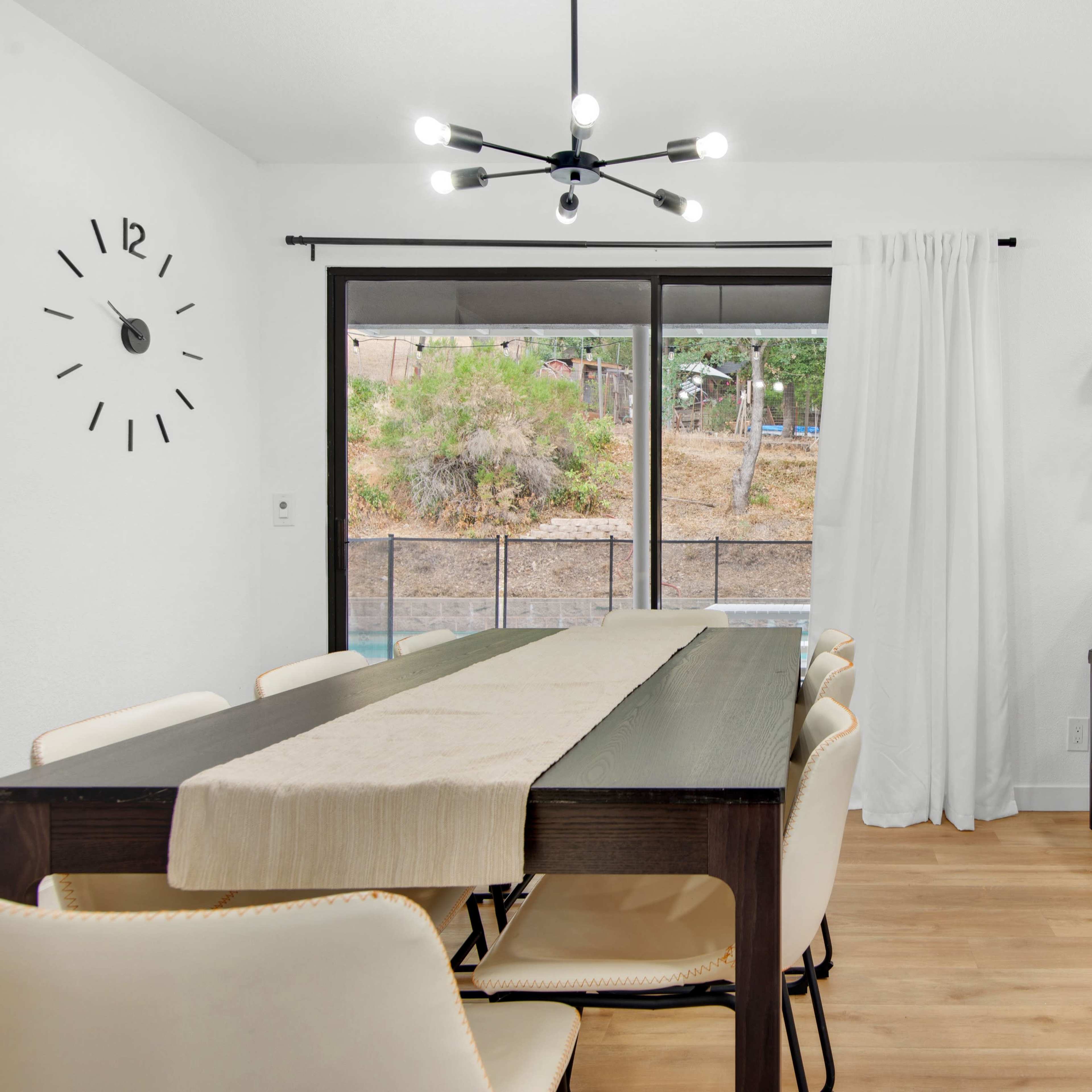 A modern dining area with a long table, chairs, and a bar counter, featuring a minimalist clock on the wall and large windows overlooking a garden.