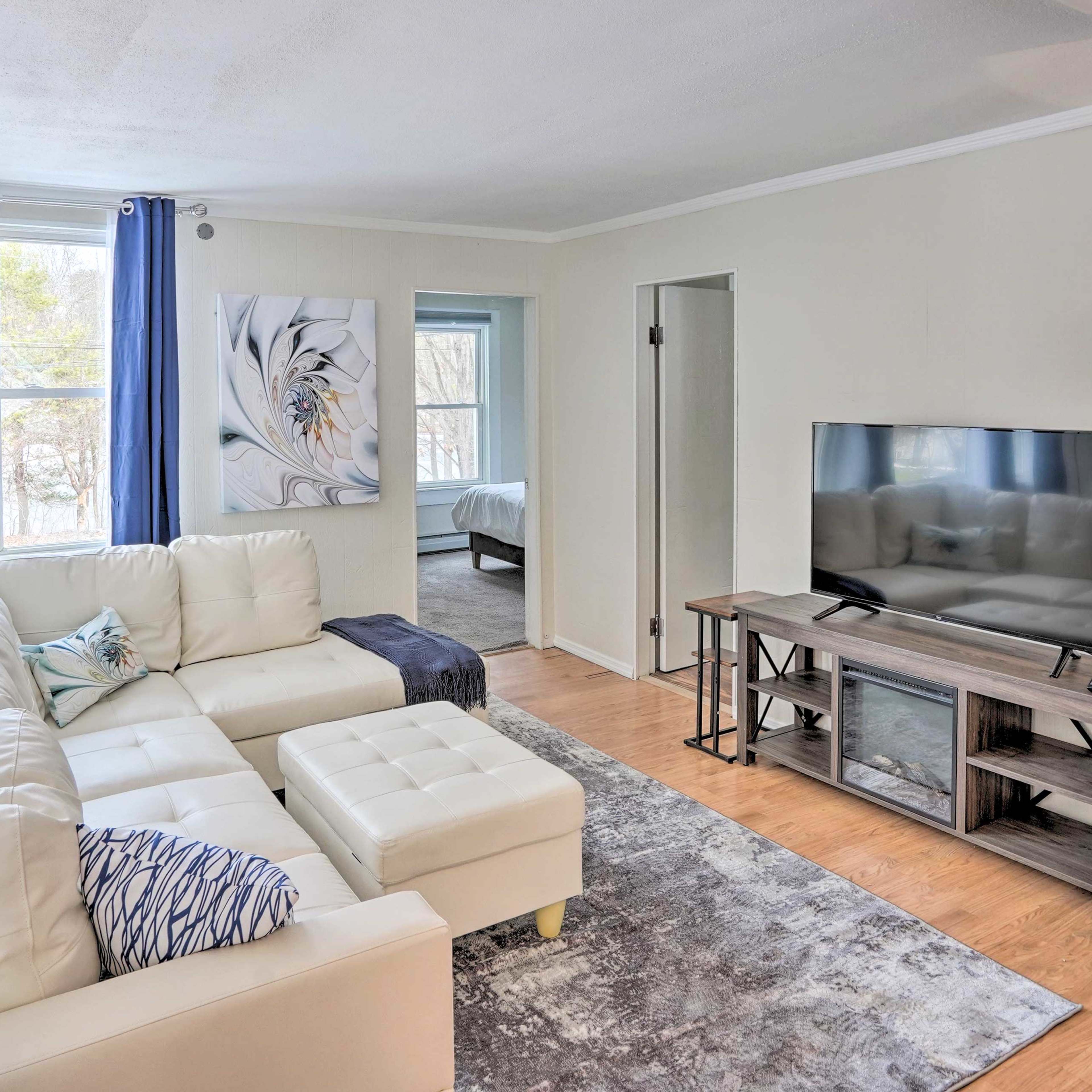 A bright living room with a white sectional sofa, a large television on a stand, a light gray rug, and blue curtains near a window.