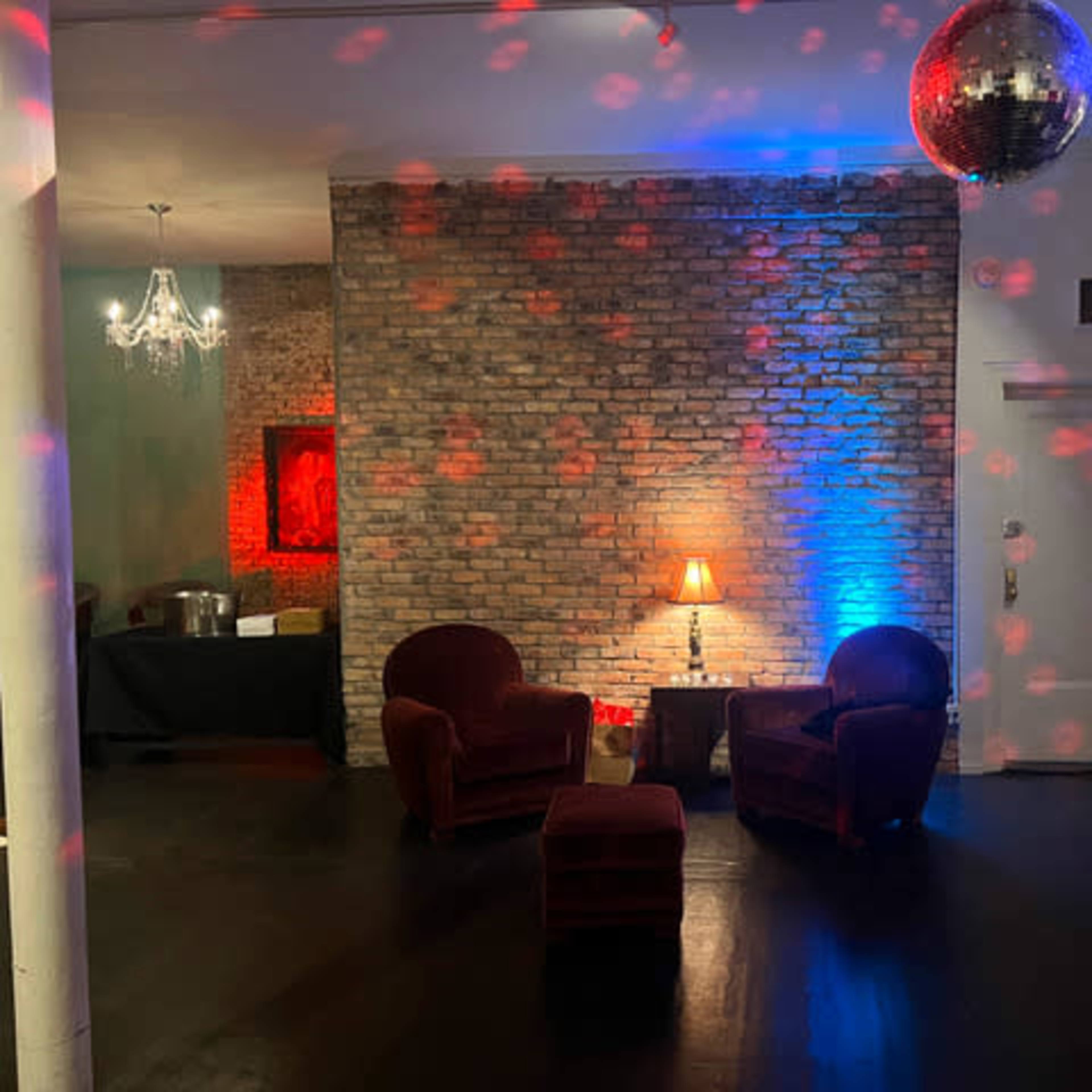 A dimly lit room with two red chairs, a table lamp, a disco ball reflecting colored lights on a brick wall, and a small seating area.