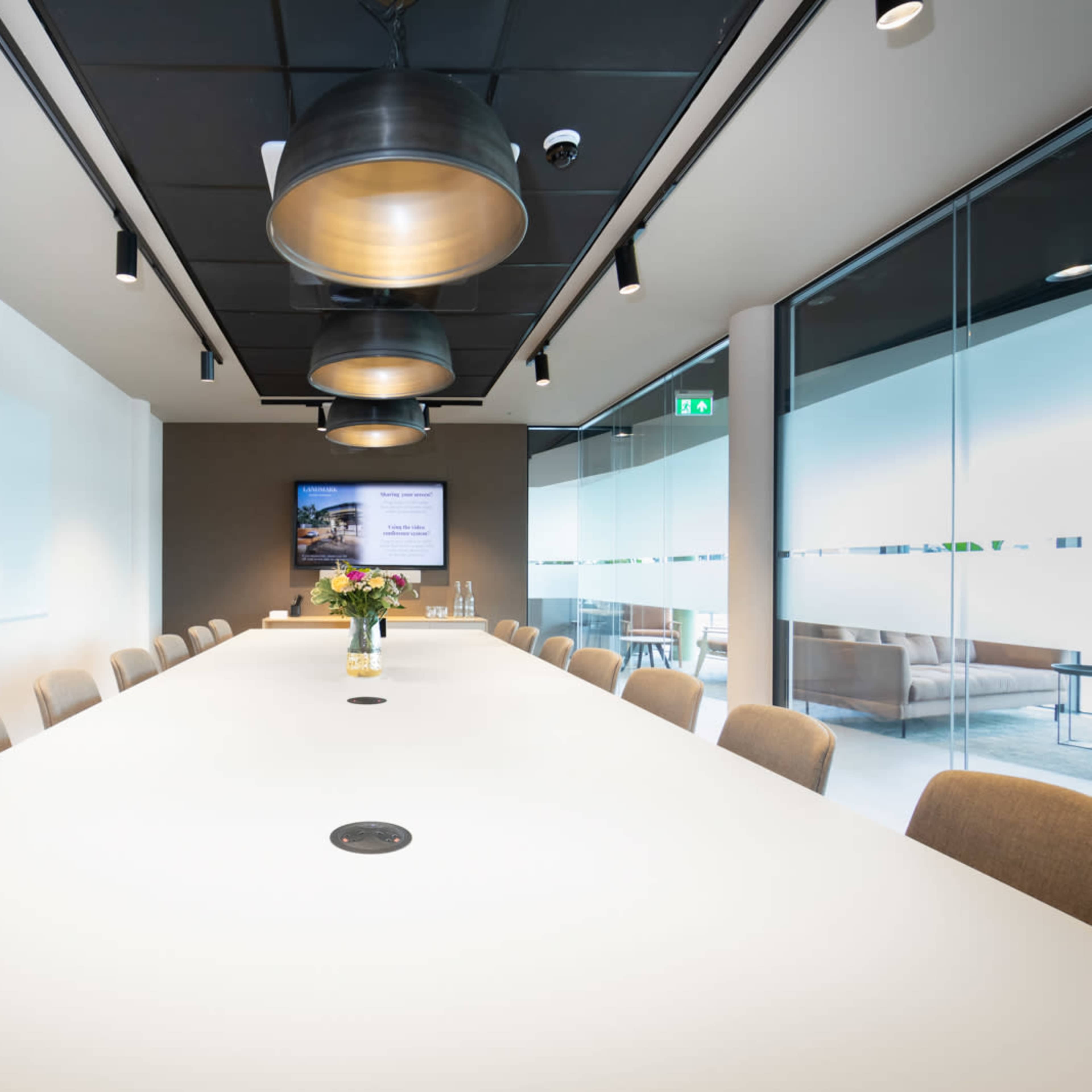 A modern conference room features a long white table surrounded by beige chairs, with pendant lighting overhead and a large screen at one end.