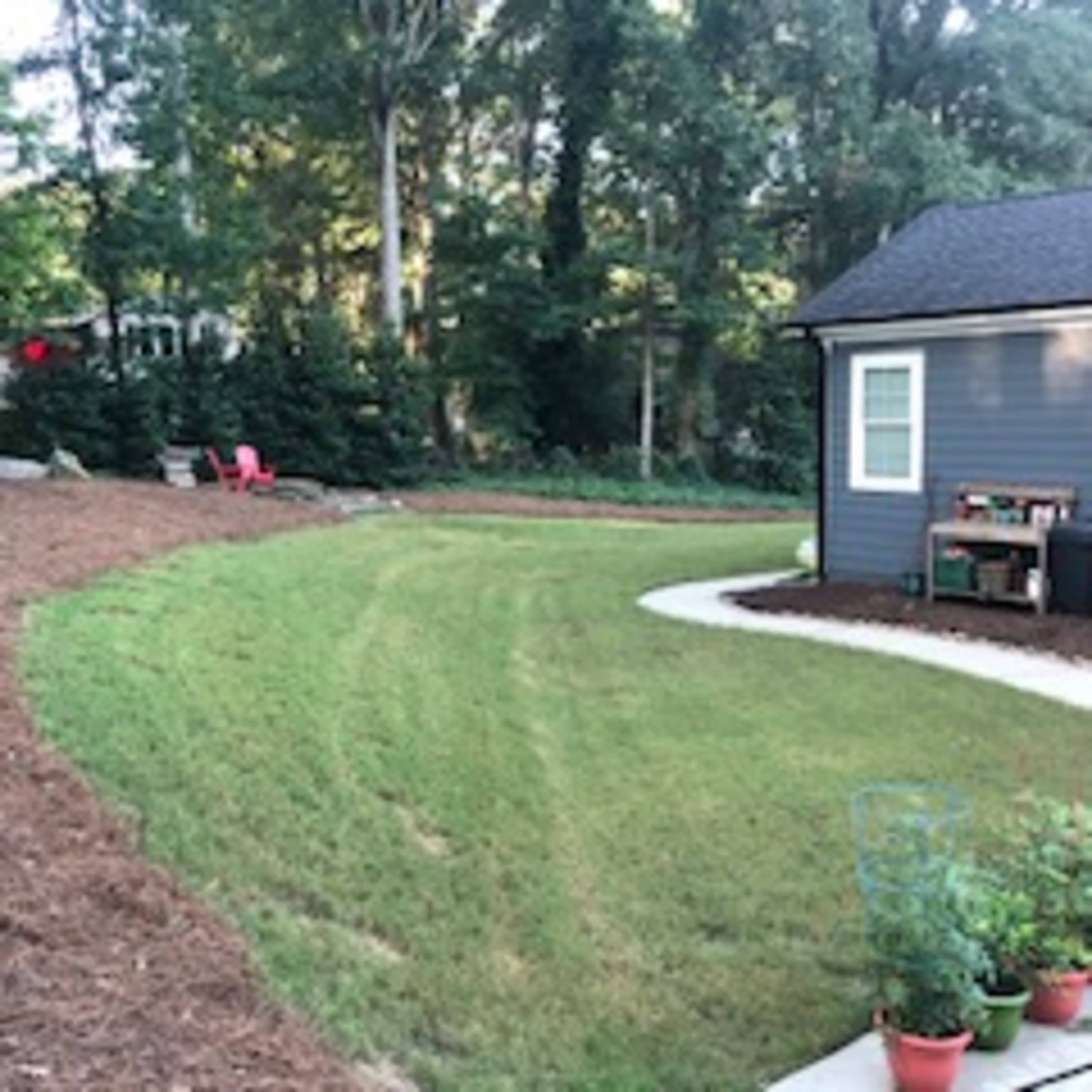 A landscaped yard featuring a curved lawn with freshly laid mulch, a pathway, and a small seating area near the house.