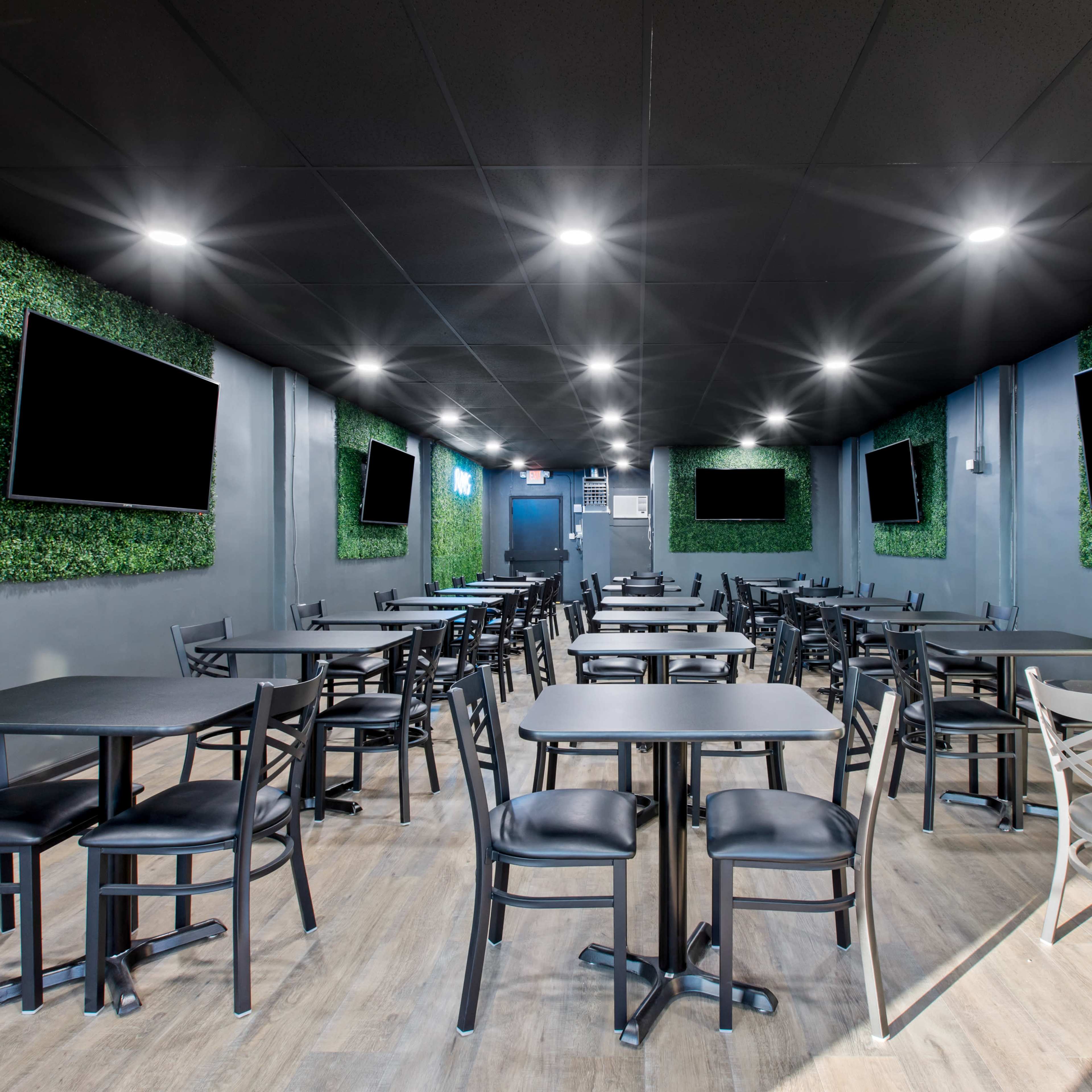 A modern dining area with multiple black tables and chairs, surrounded by green wall panels and equipped with wall-mounted televisions.