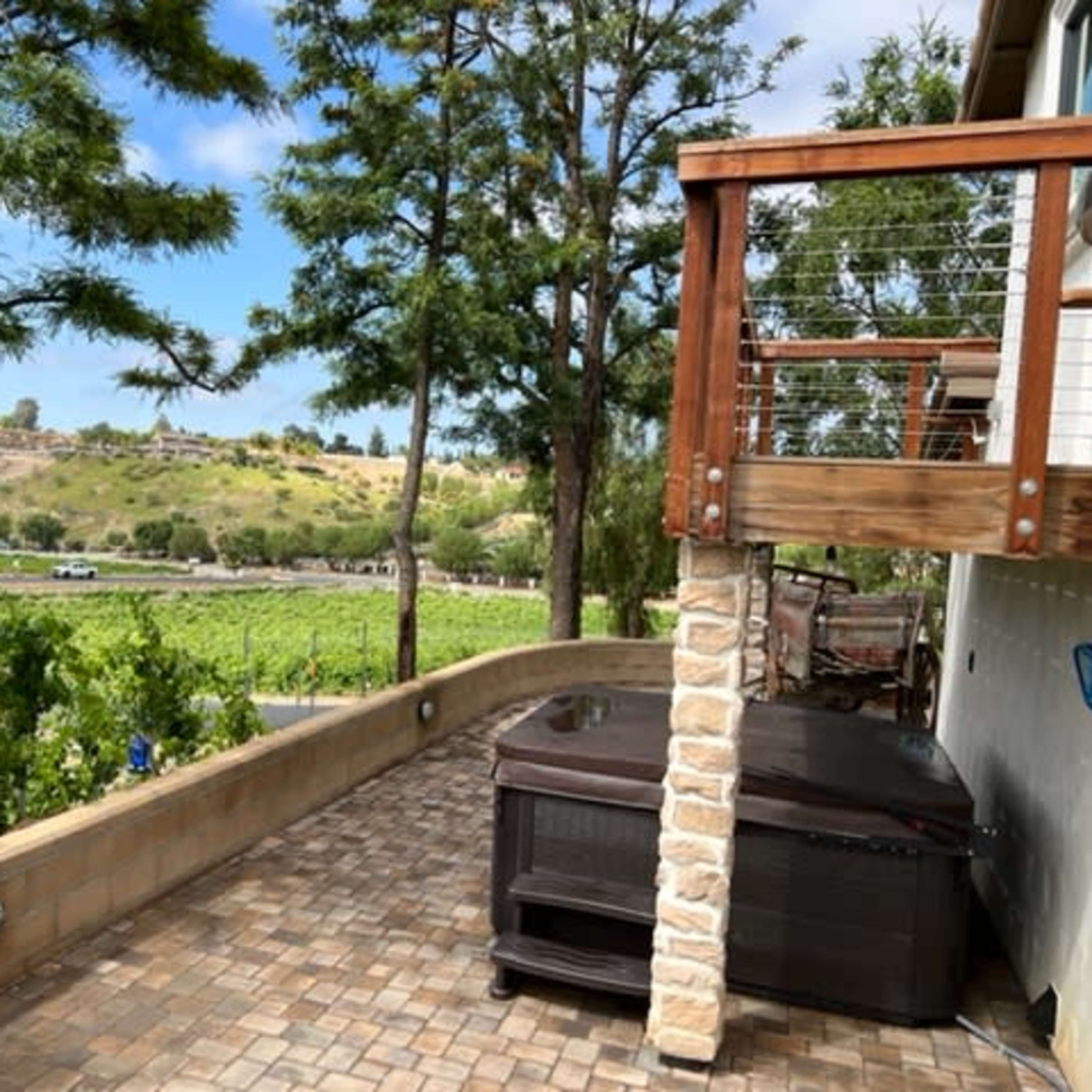The scene shows a patio with a hot tub, surrounded by trees, overlooking a vineyard and a road in the distance.