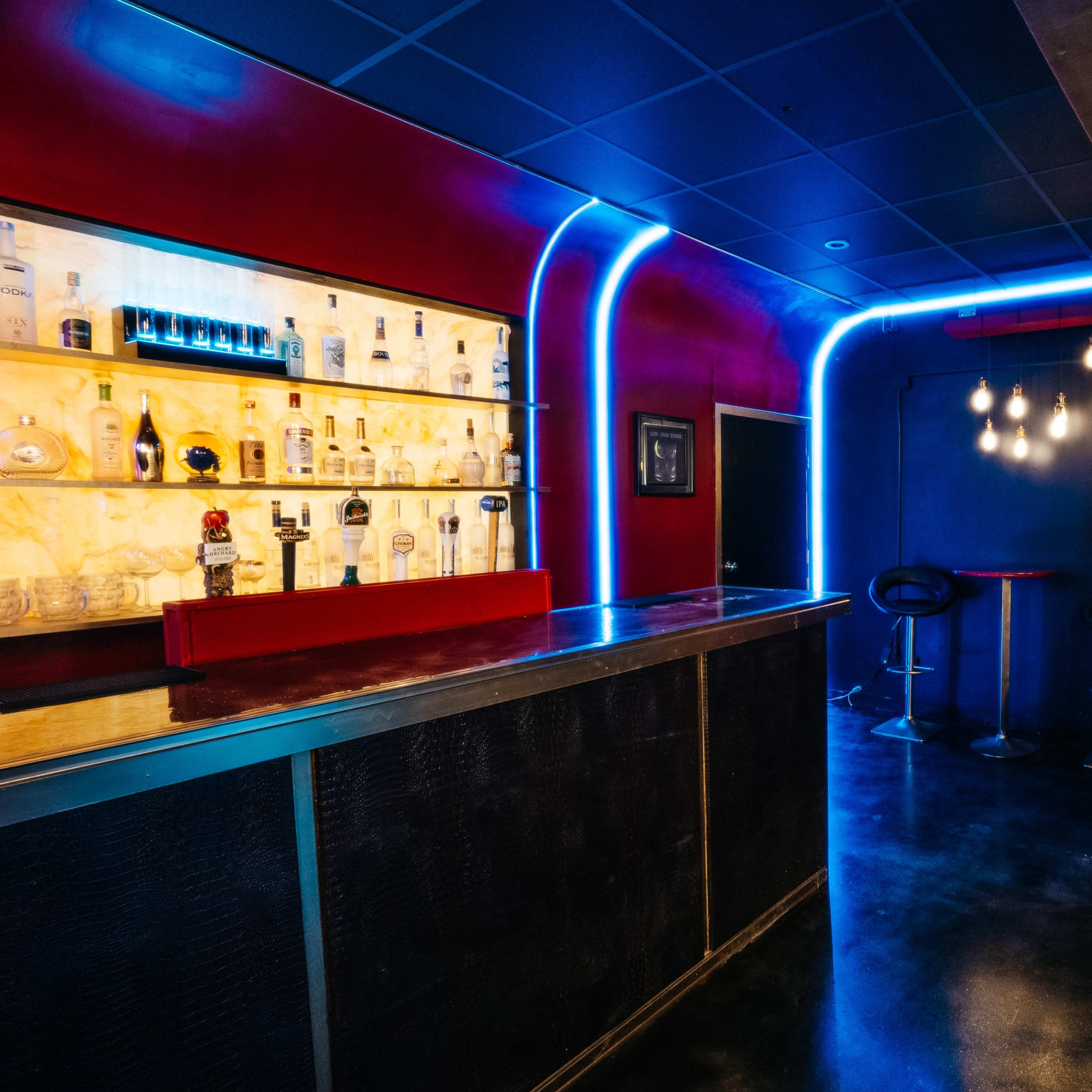 A modern bar area with a brightly lit backlit shelf displaying various bottles and seating along the walls.