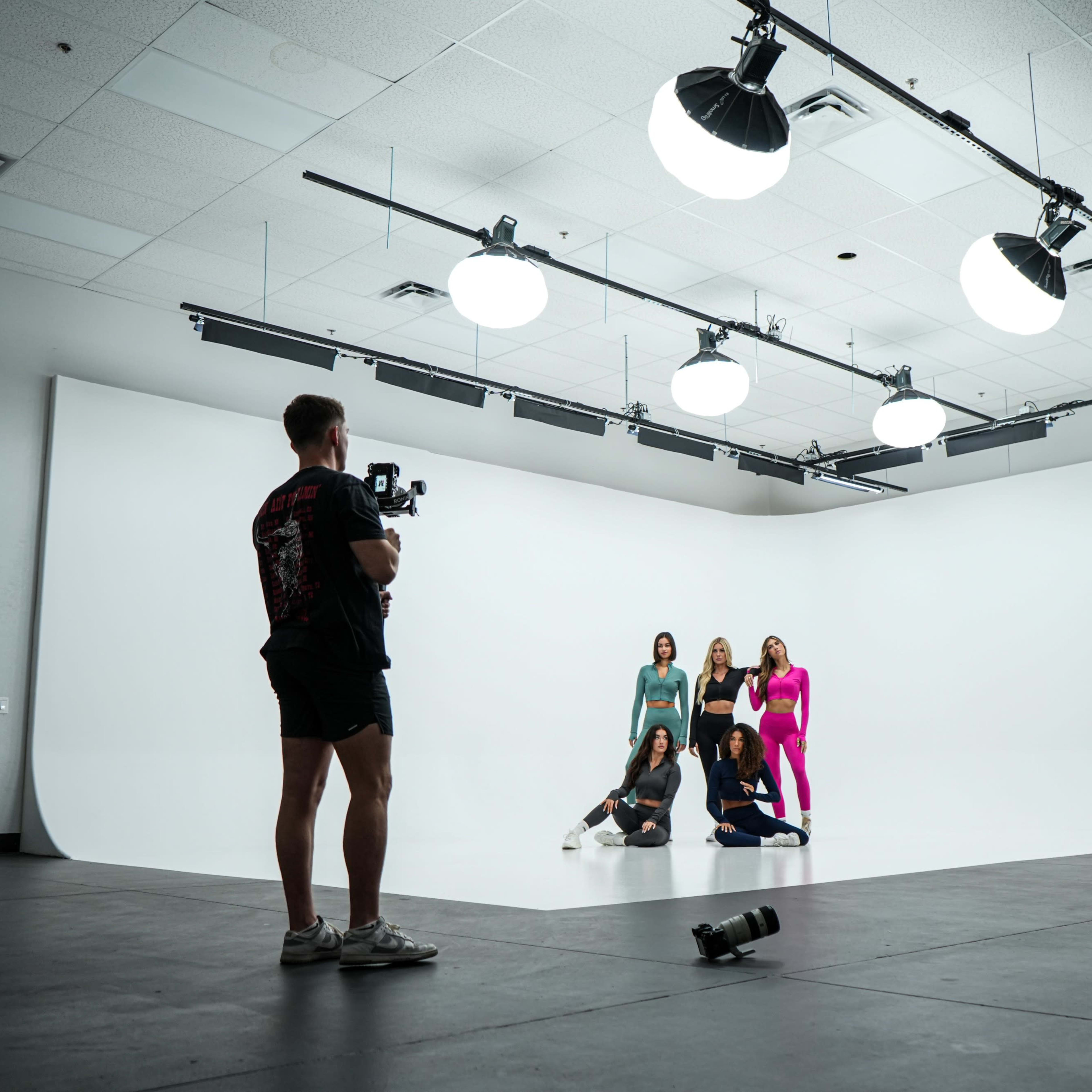 A photographer captures a group of models posing on a white backdrop while studio lights illuminate the scene.