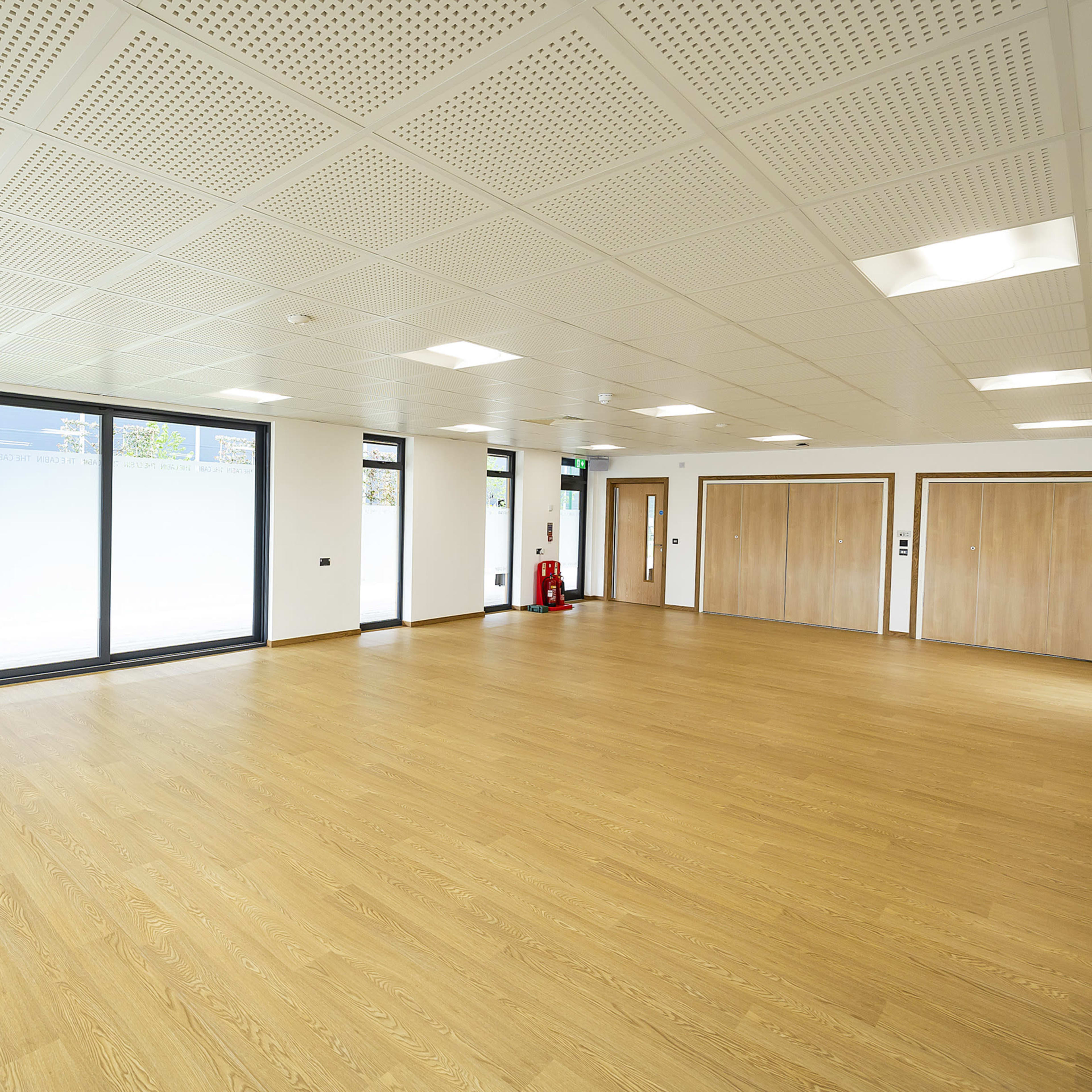 An empty indoor space with wooden flooring, large windows, and two closed doors on one side.
