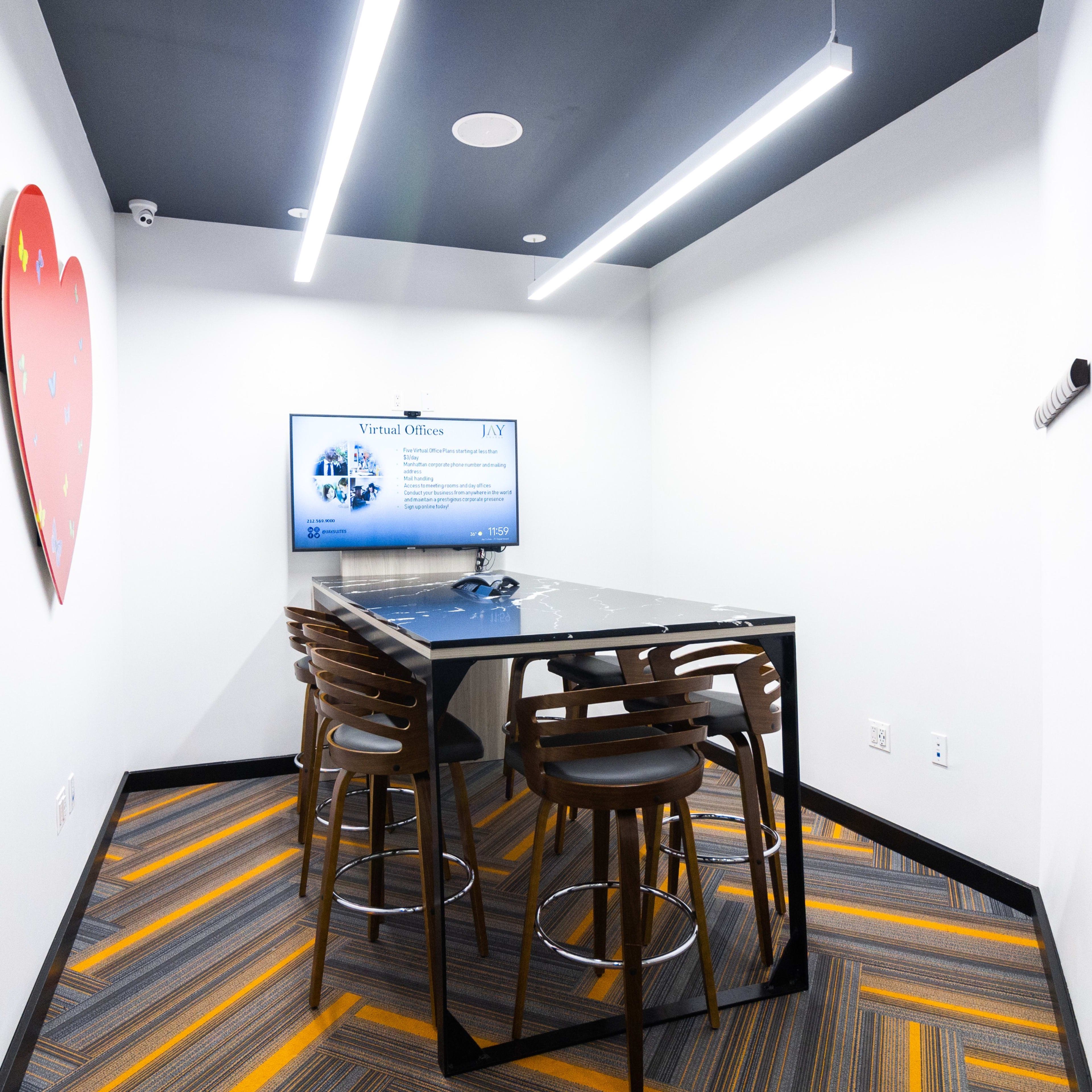 A modern meeting room features a glass table surrounded by high stools, a television screen displaying a presentation, and a heart-shaped decoration on the wall.