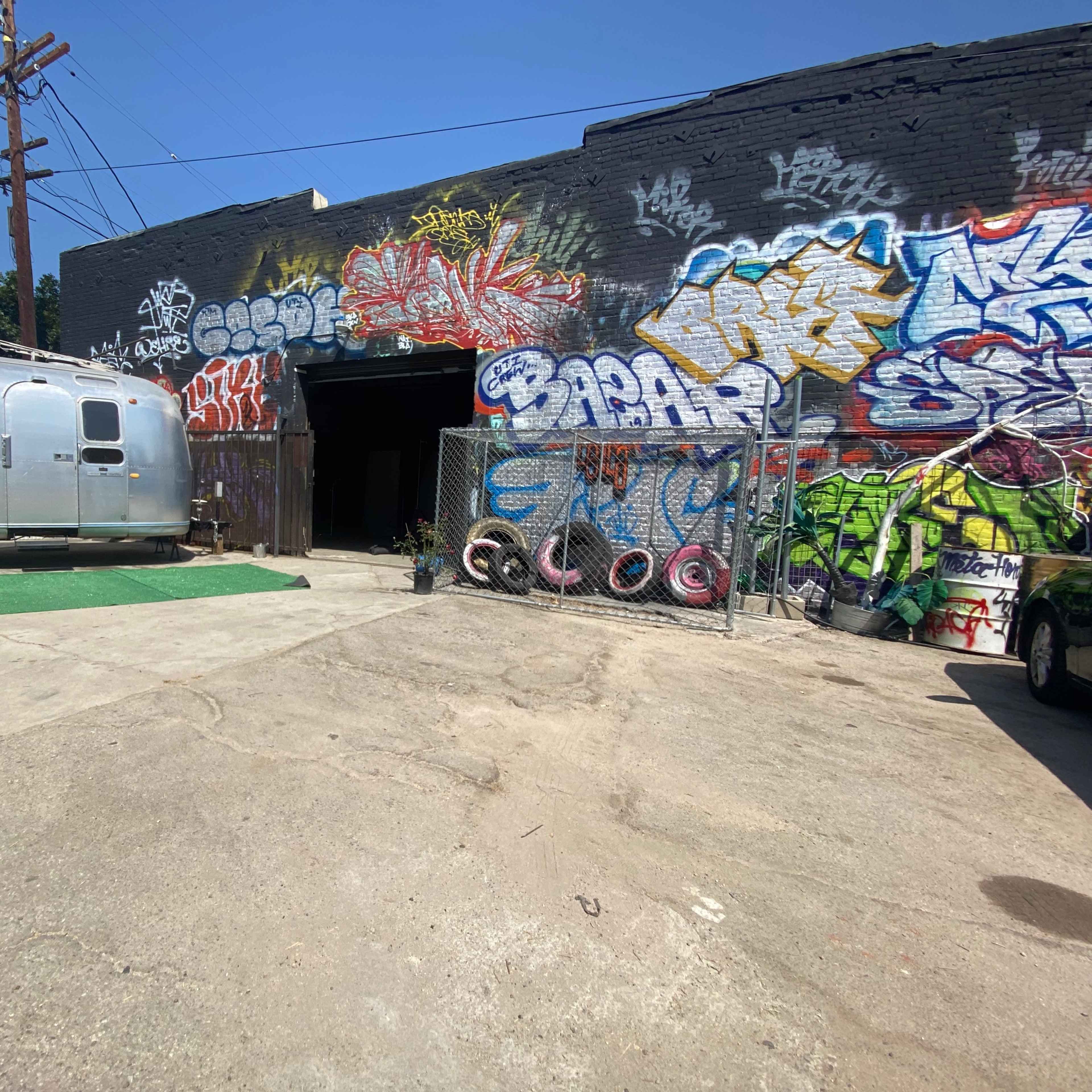 An Airstream trailer is parked next to a graffiti-covered warehouse entrance on a concrete lot.