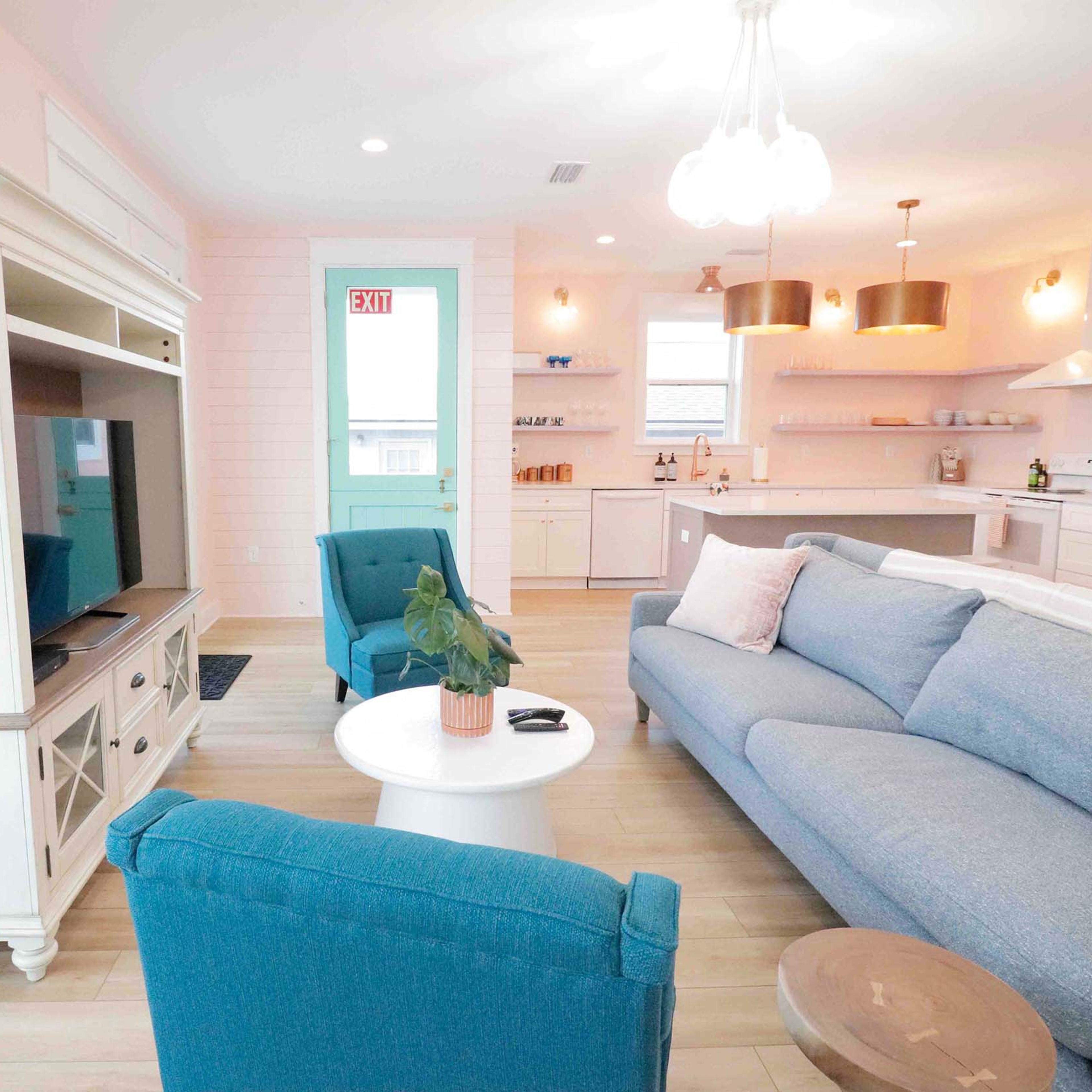 A brightly lit living room with a blue couch, turquoise chairs, a white coffee table, and a modern kitchen in the background.