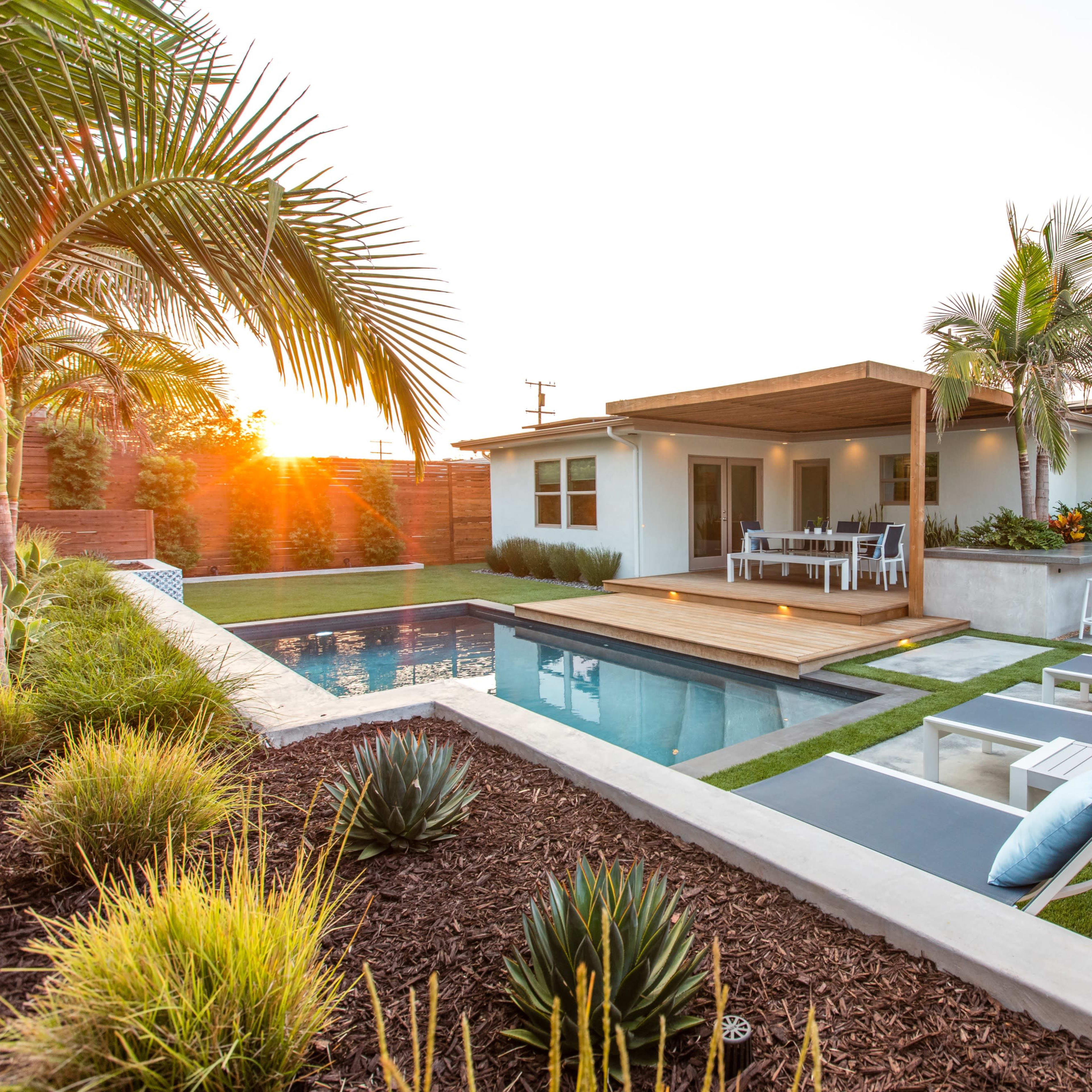 A modern backyard with a swimming pool, wooden deck, and seating area, surrounded by landscaping and palm trees at sunset.