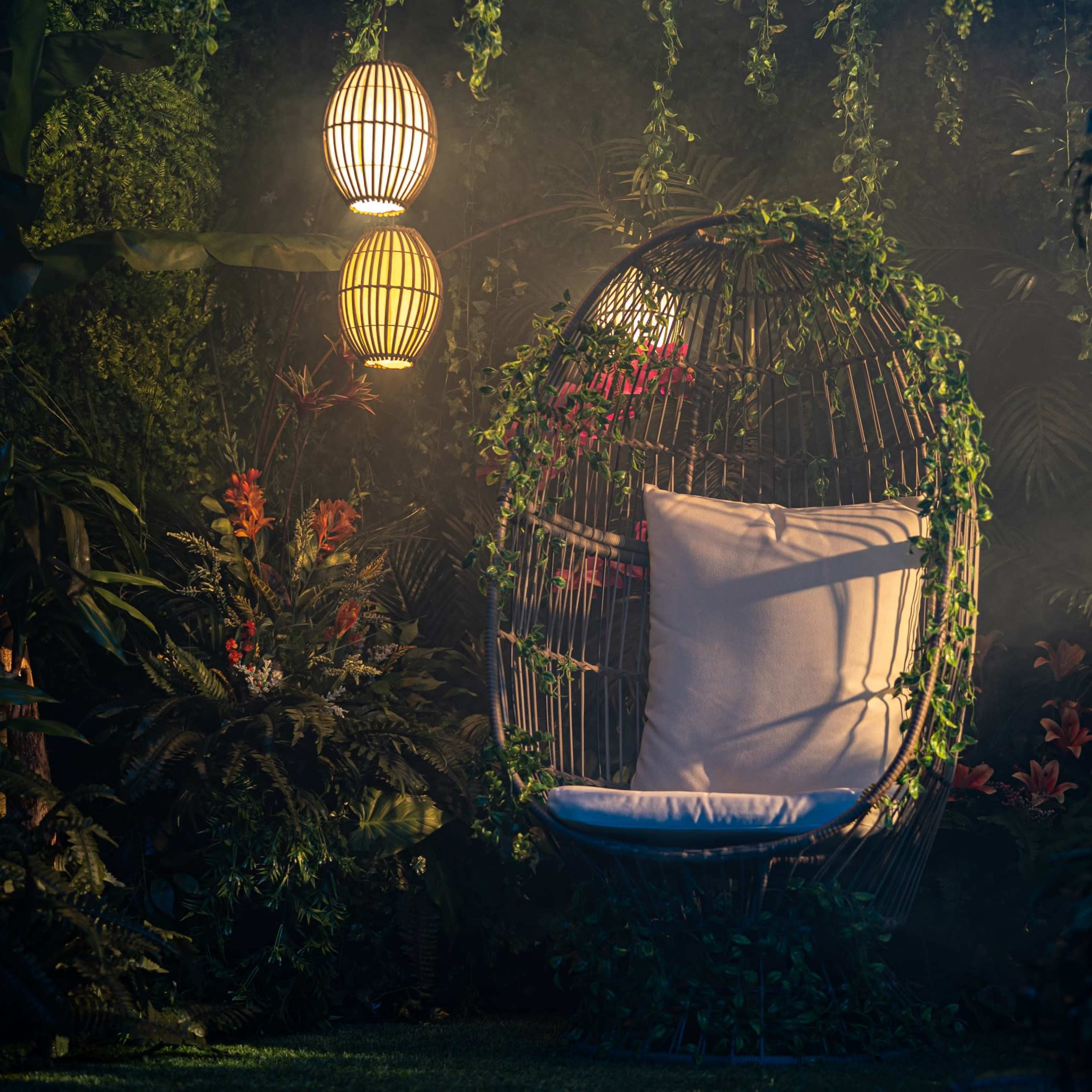 A suspended wicker chair surrounded by lush greenery and softly glowing lanterns creates a tranquil outdoor setting.