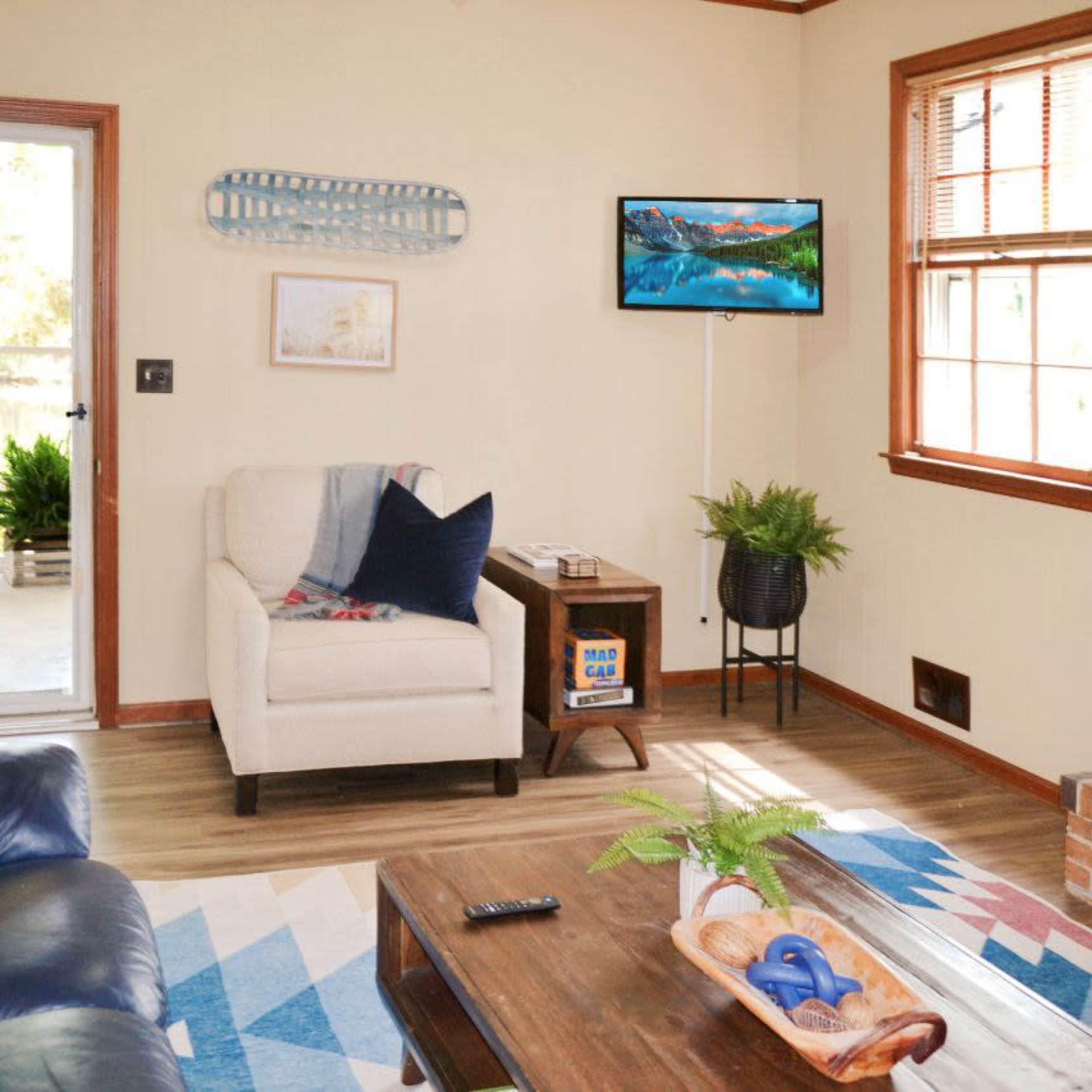 A cozy living room with a beige armchair, a blue couch, a wooden coffee table, a television mounted on the wall, and a view of a door leading outside.