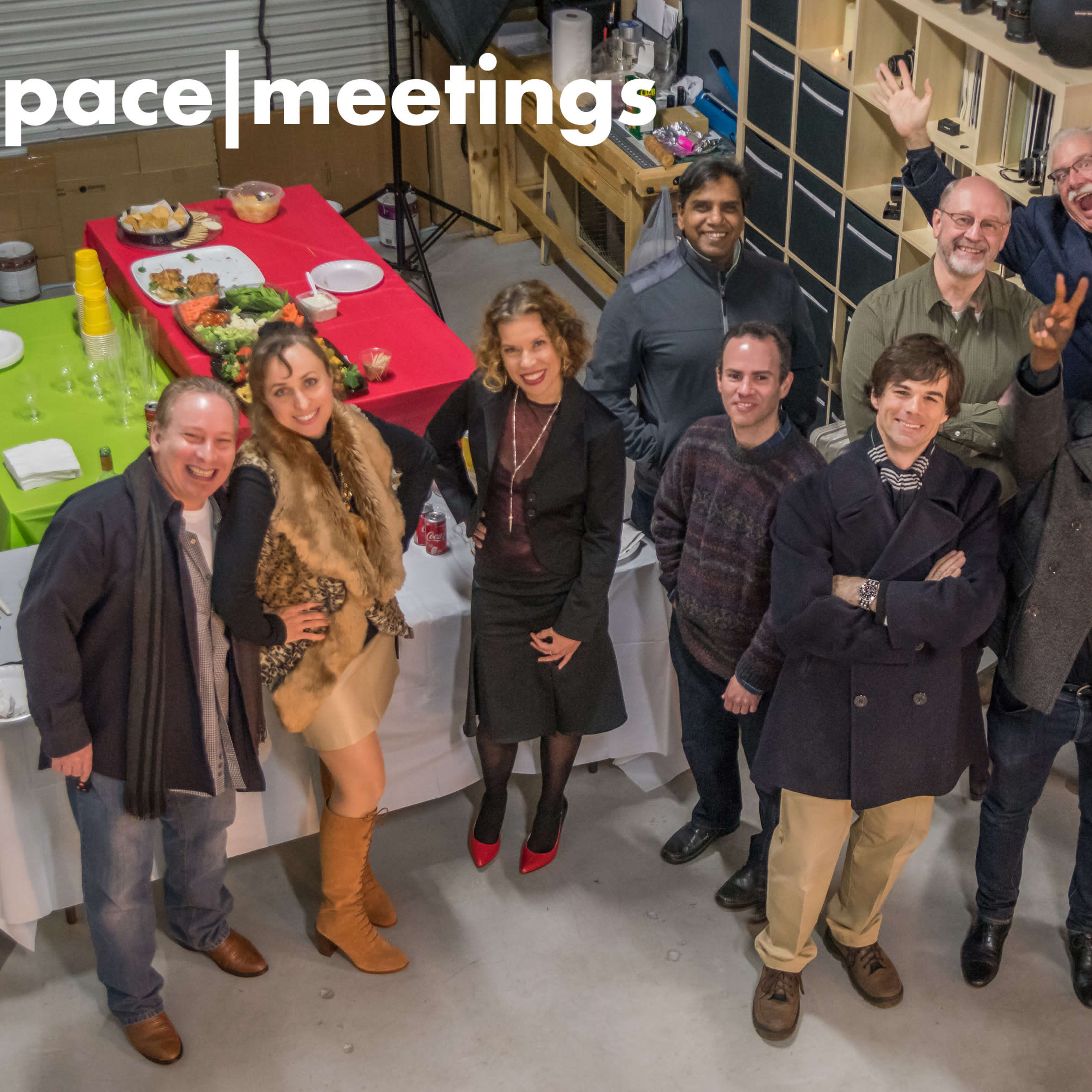 A group of twelve people is gathered in a warehouse, smiling and posing for the camera with food and drinks on a table in the background.