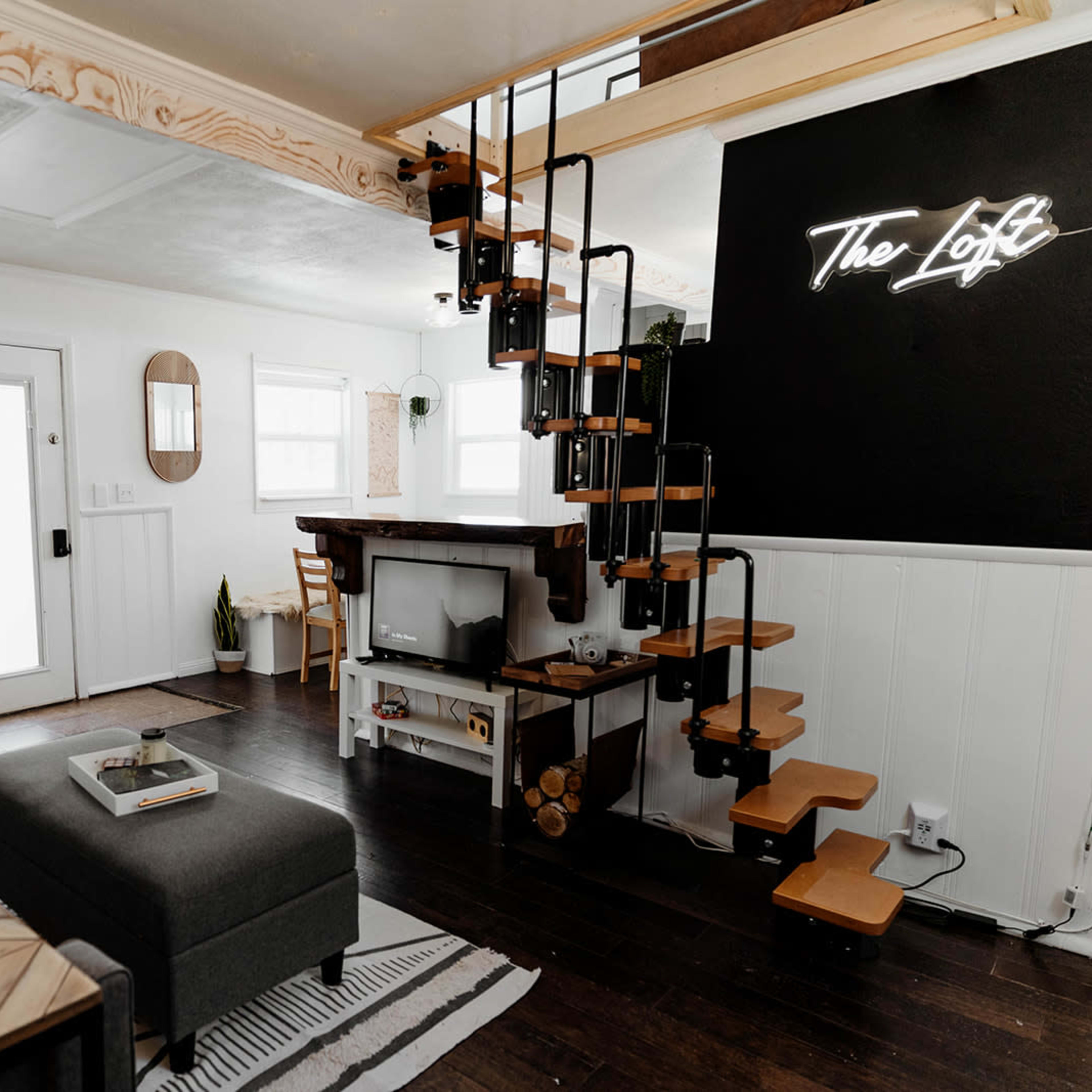 A modern living space featuring a cozy seating area, a staircase leading to a loft, and a black wall with a neon sign that reads "The Loft."