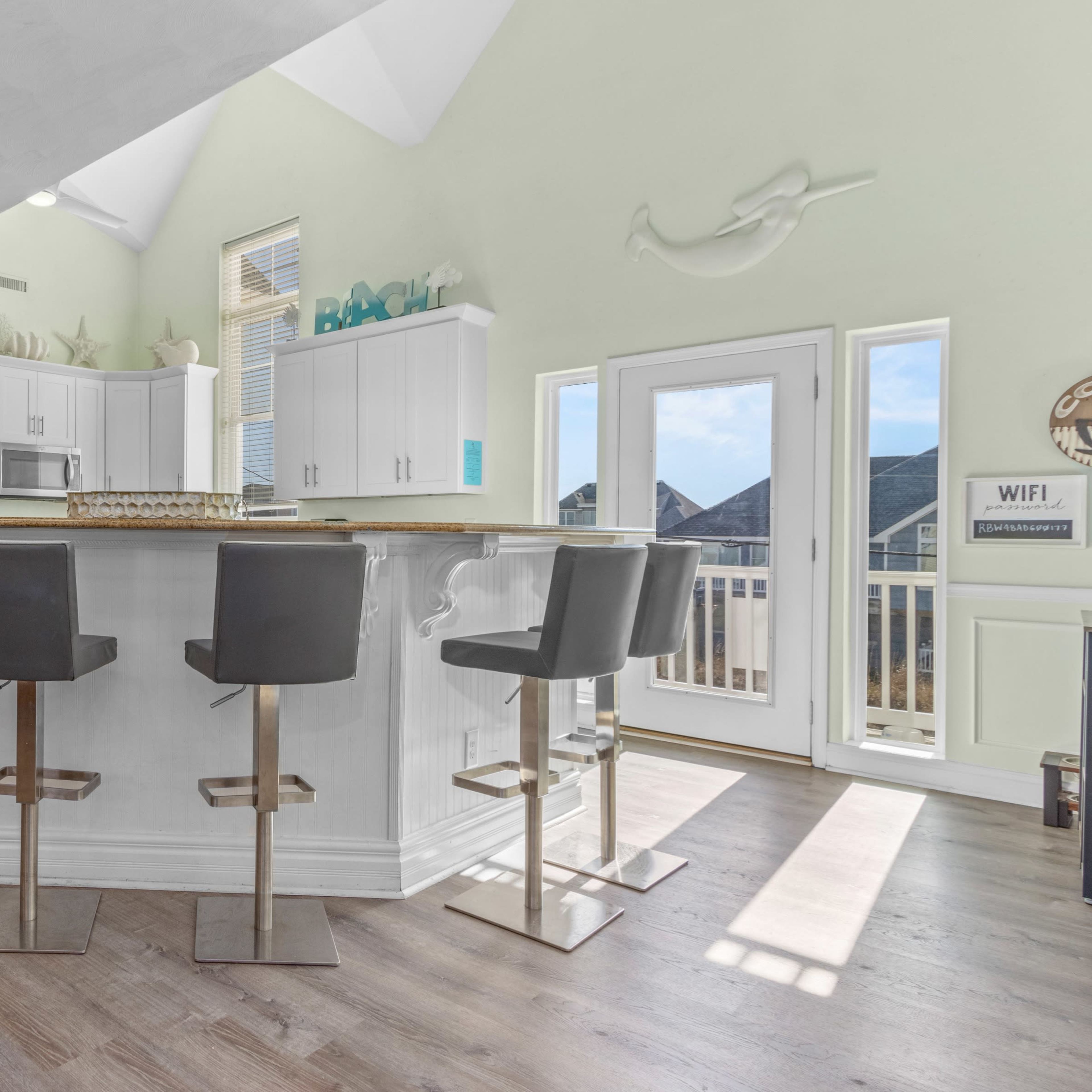A modern kitchen features a central island with high-backed chairs, white cabinetry, and large windows allowing natural light to fill the space.