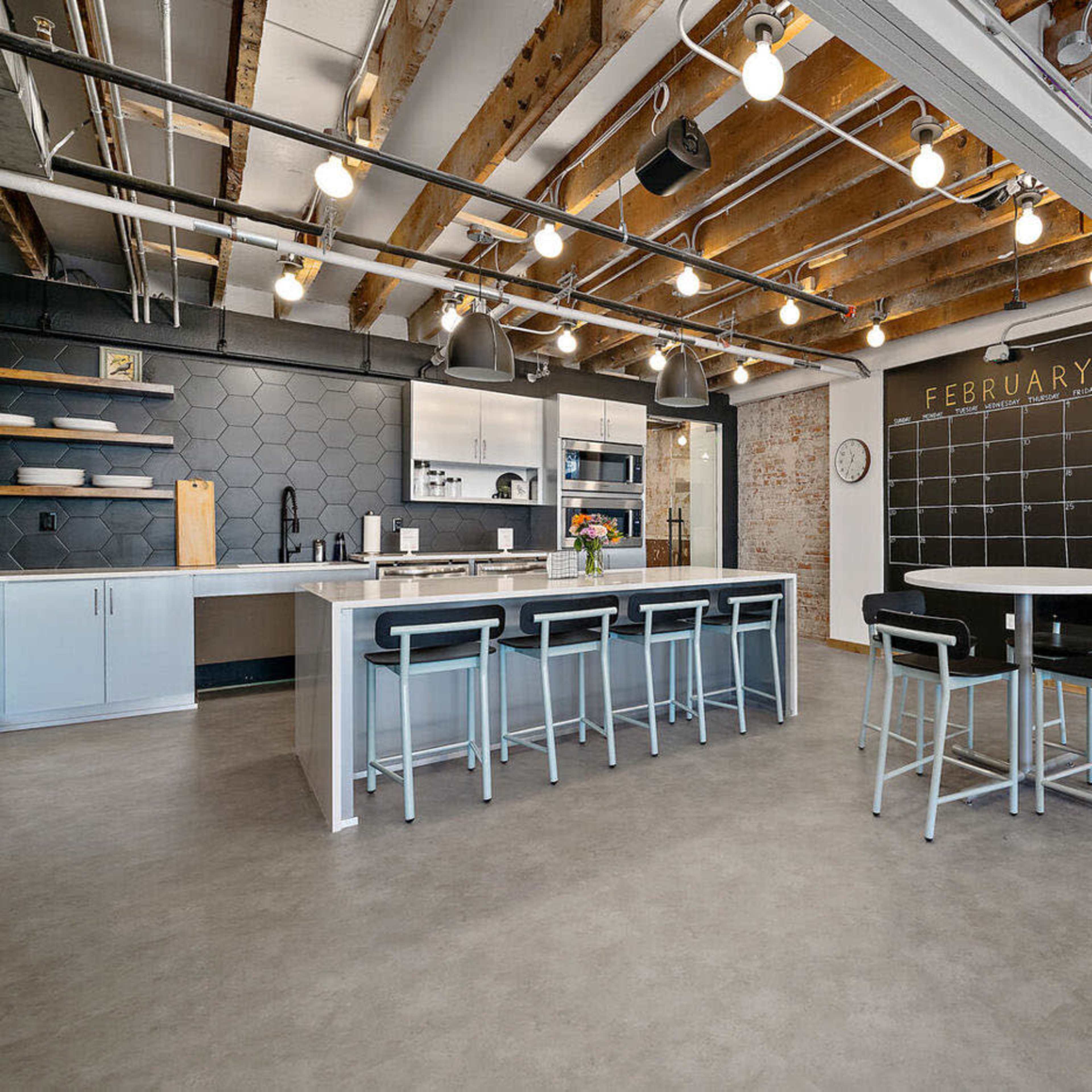 A modern kitchen and dining area with exposed beams, a large island, bar stools, and a chalkboard wall displaying the month of February.