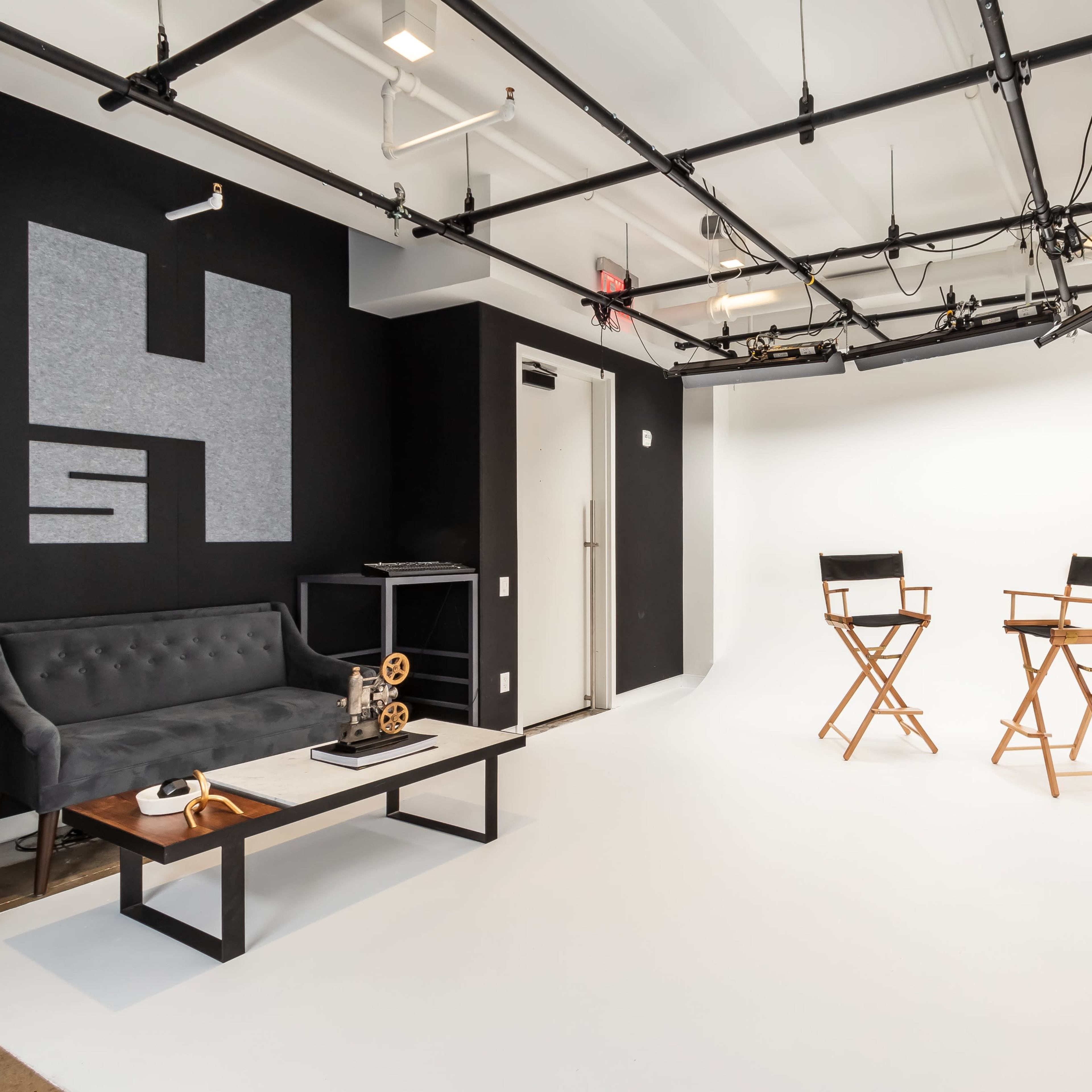 A minimalist studio space featuring two director's chairs in front of a white backdrop, accompanied by a black couch and a coffee table.