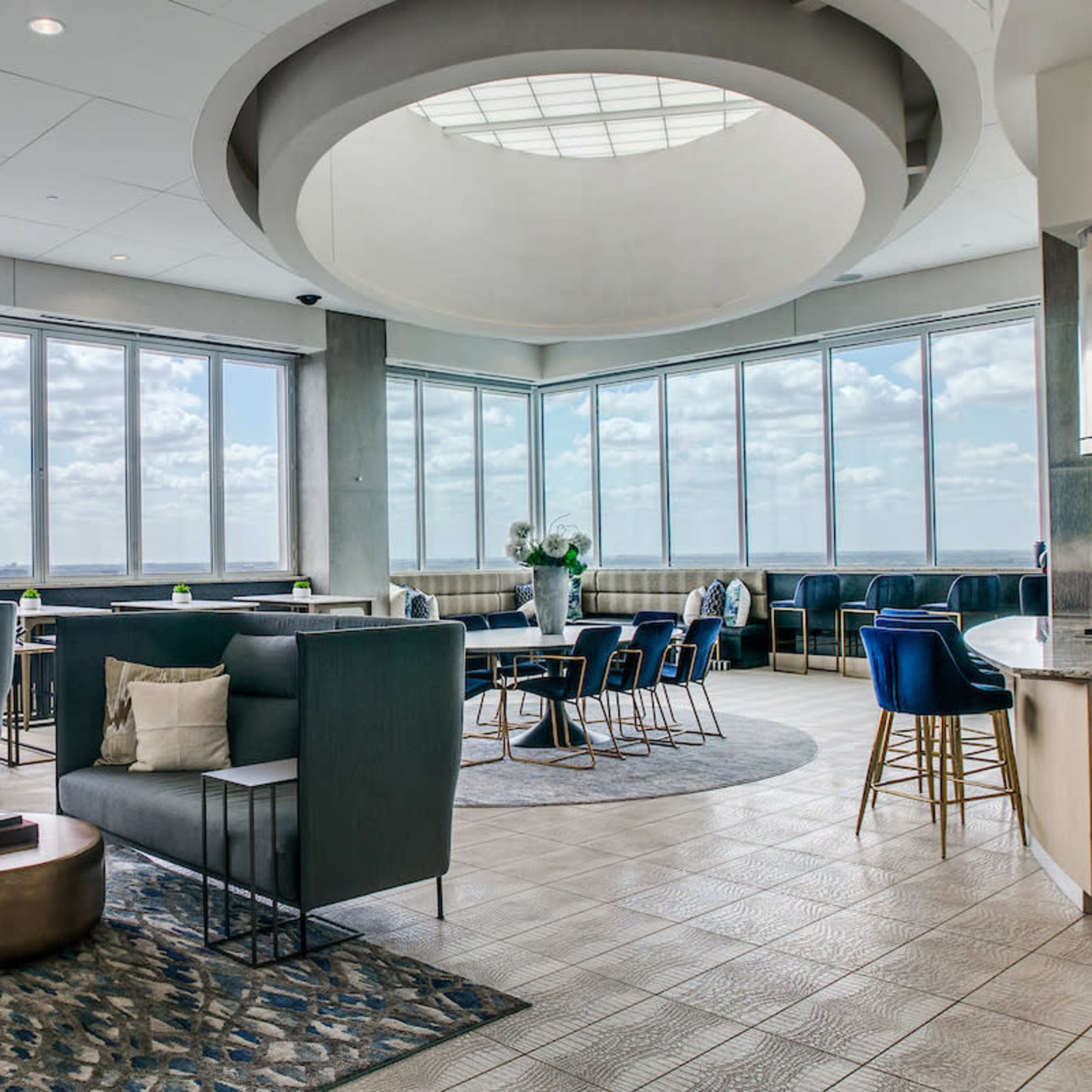A modern lounge area features large windows with a view, comfortable seating, and decorative rugs.