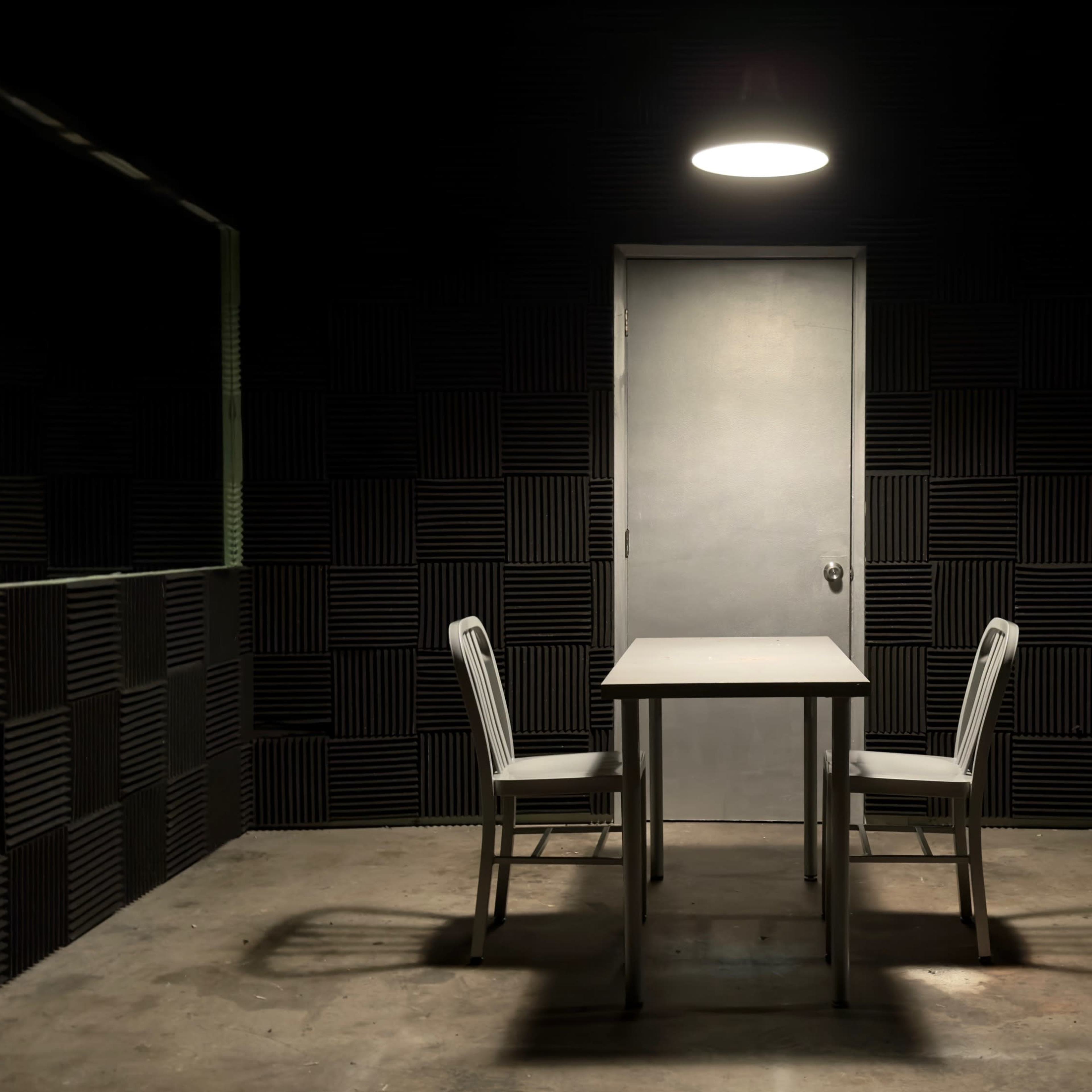 A minimal interrogation room features a table with two chairs and a single overhead light, surrounded by soundproofed walls and mirrors.