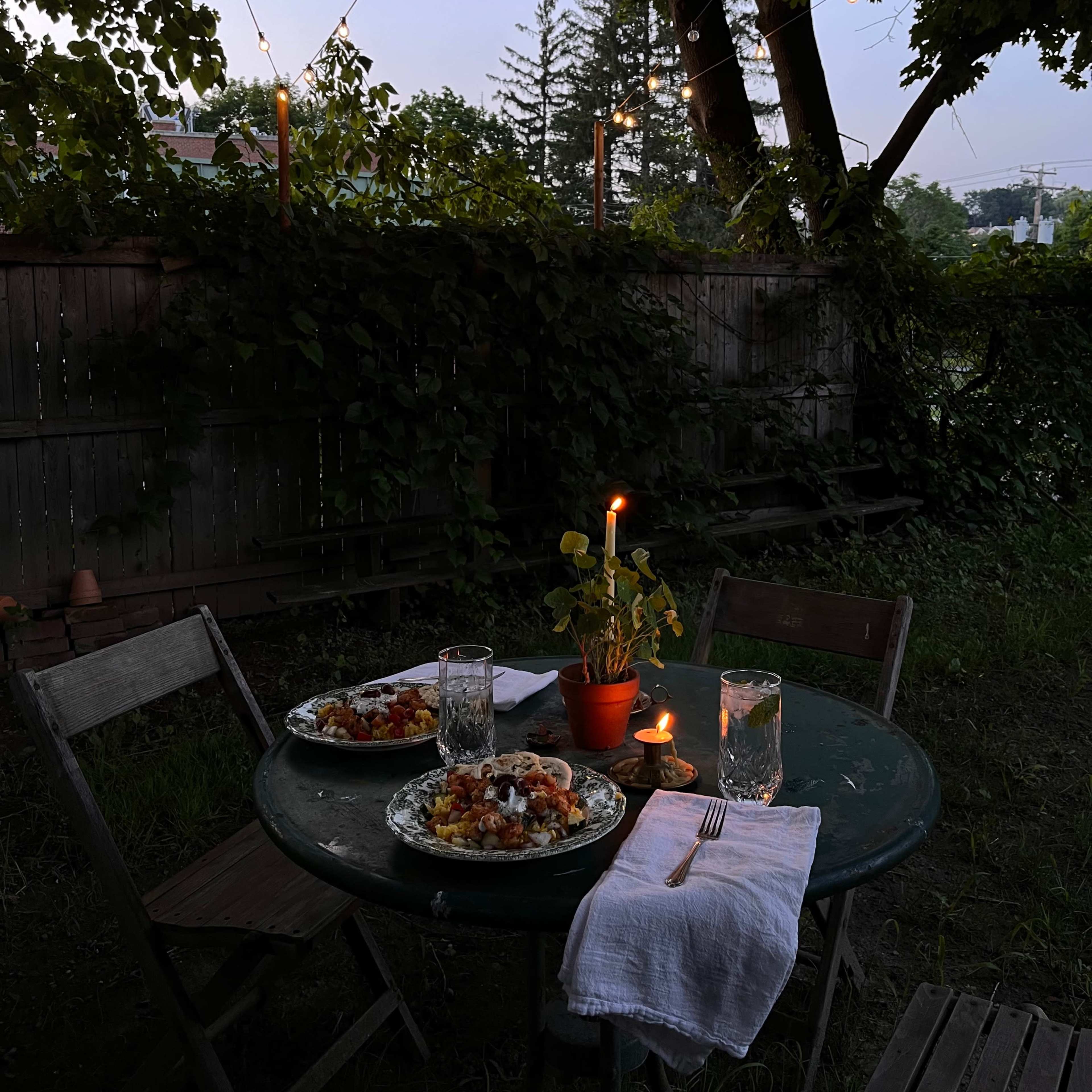 A small round table is set for two with plates of food, glasses of water, and a candle in a potted plant, beneath string lights illuminating a backyard at dusk.