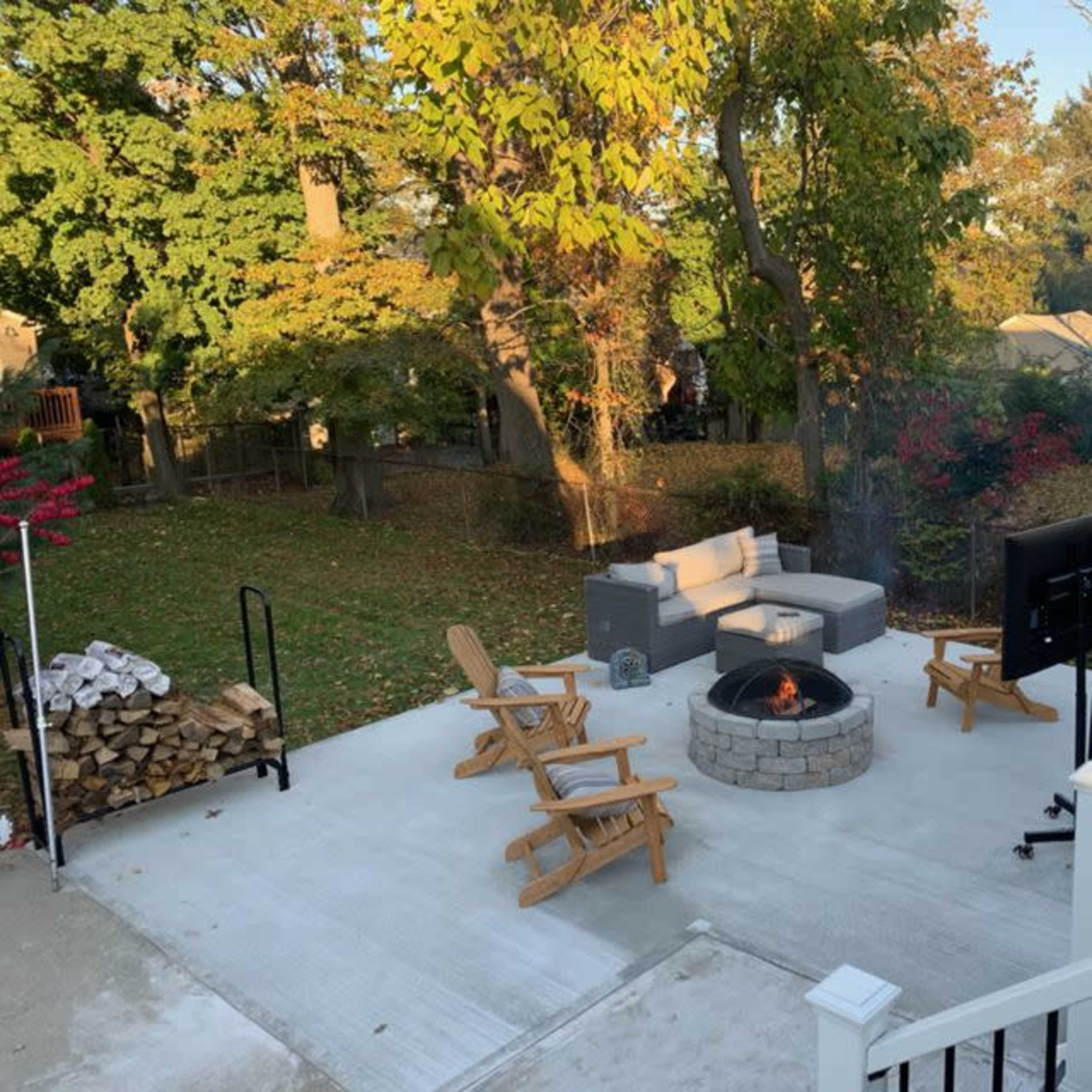 A backyard patio with a fire pit surrounded by cushioned seating and wooden chairs, framed by trees and a stack of firewood.