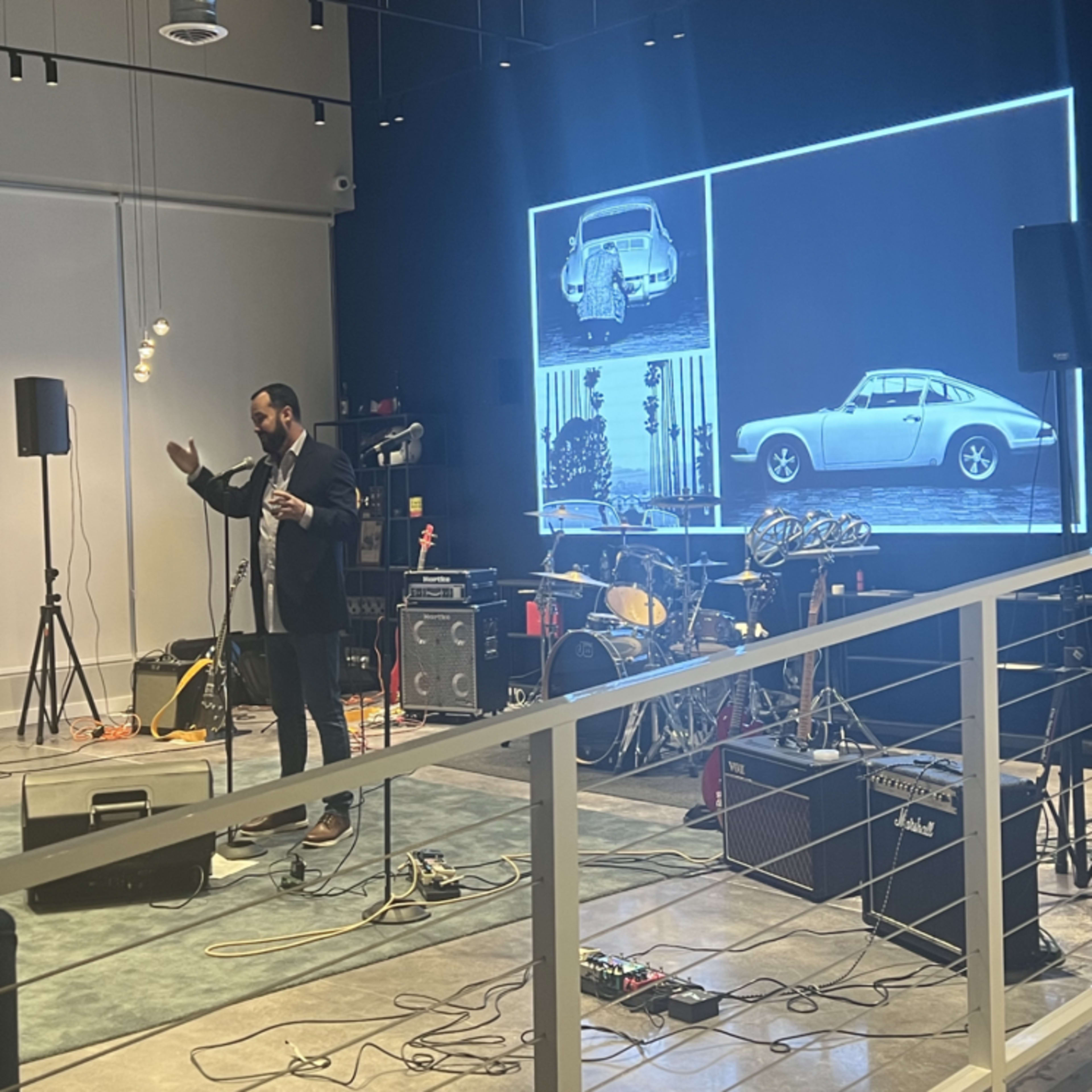 A speaker presents in front of a large screen displaying classic cars, with musical equipment set up nearby.