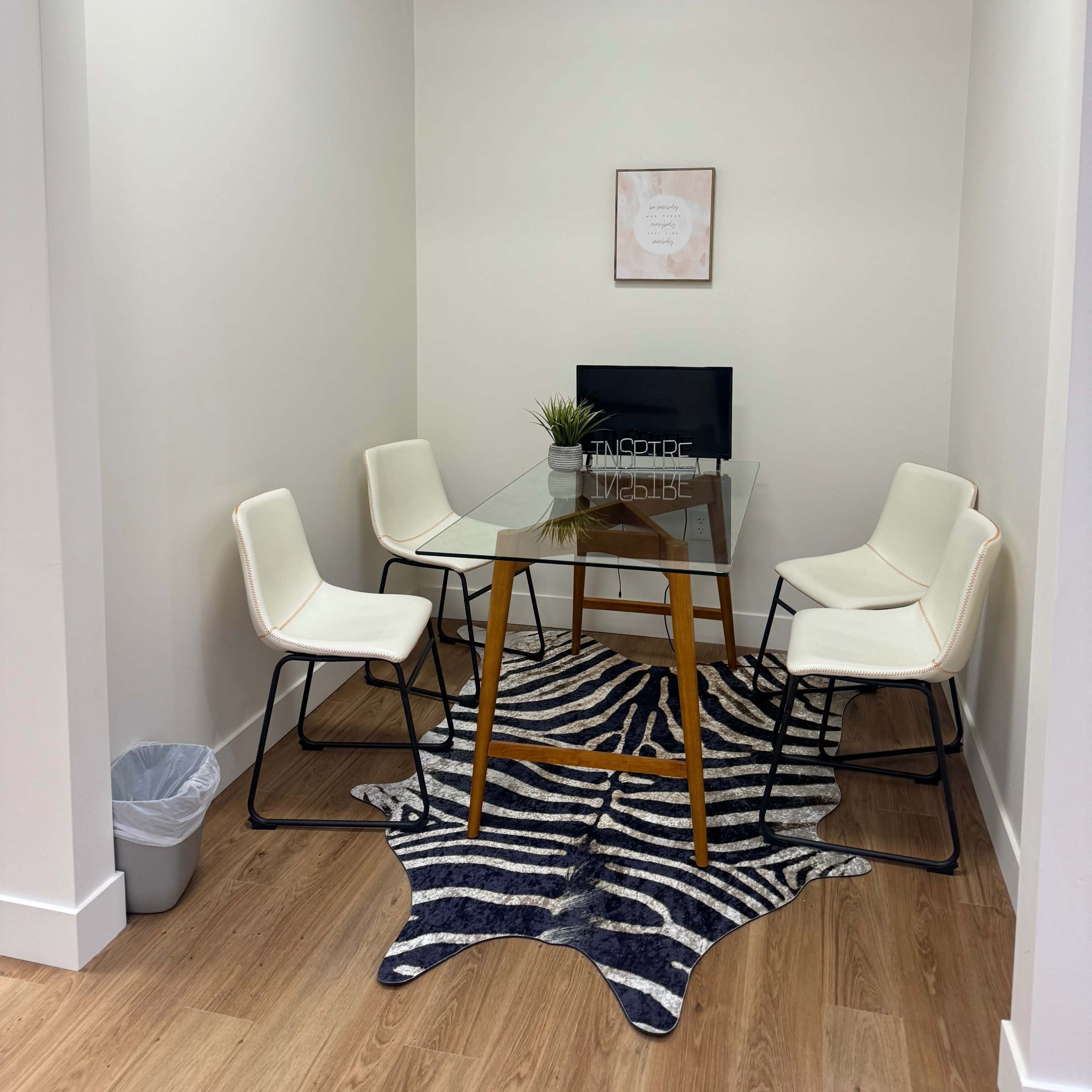 A small meeting room features a glass table surrounded by four white chairs on a zebra-striped rug, with a potted plant and a piece of wall art in the background.