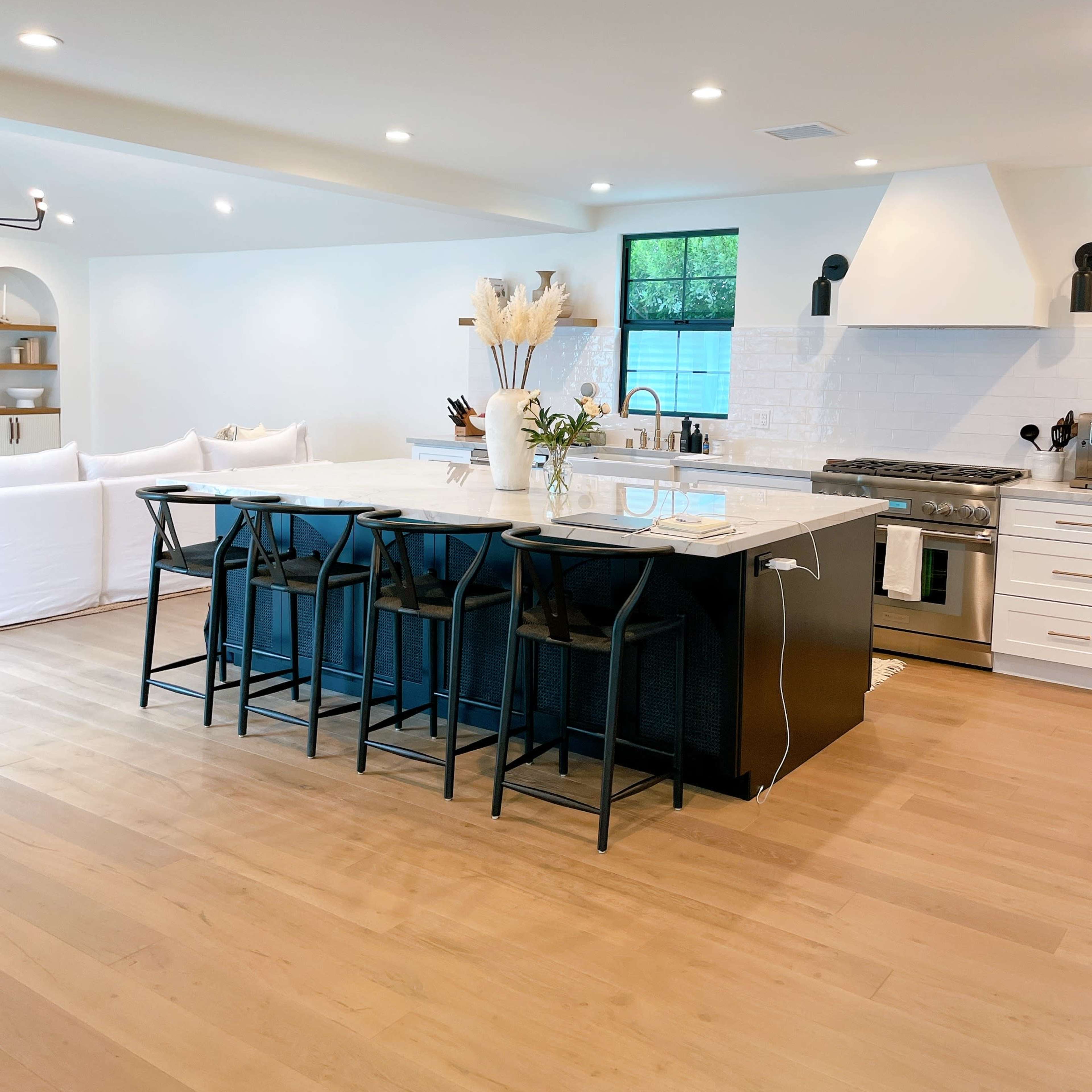 A modern kitchen features a large island with black chairs, white cabinets, and a stainless steel stove against a light background.