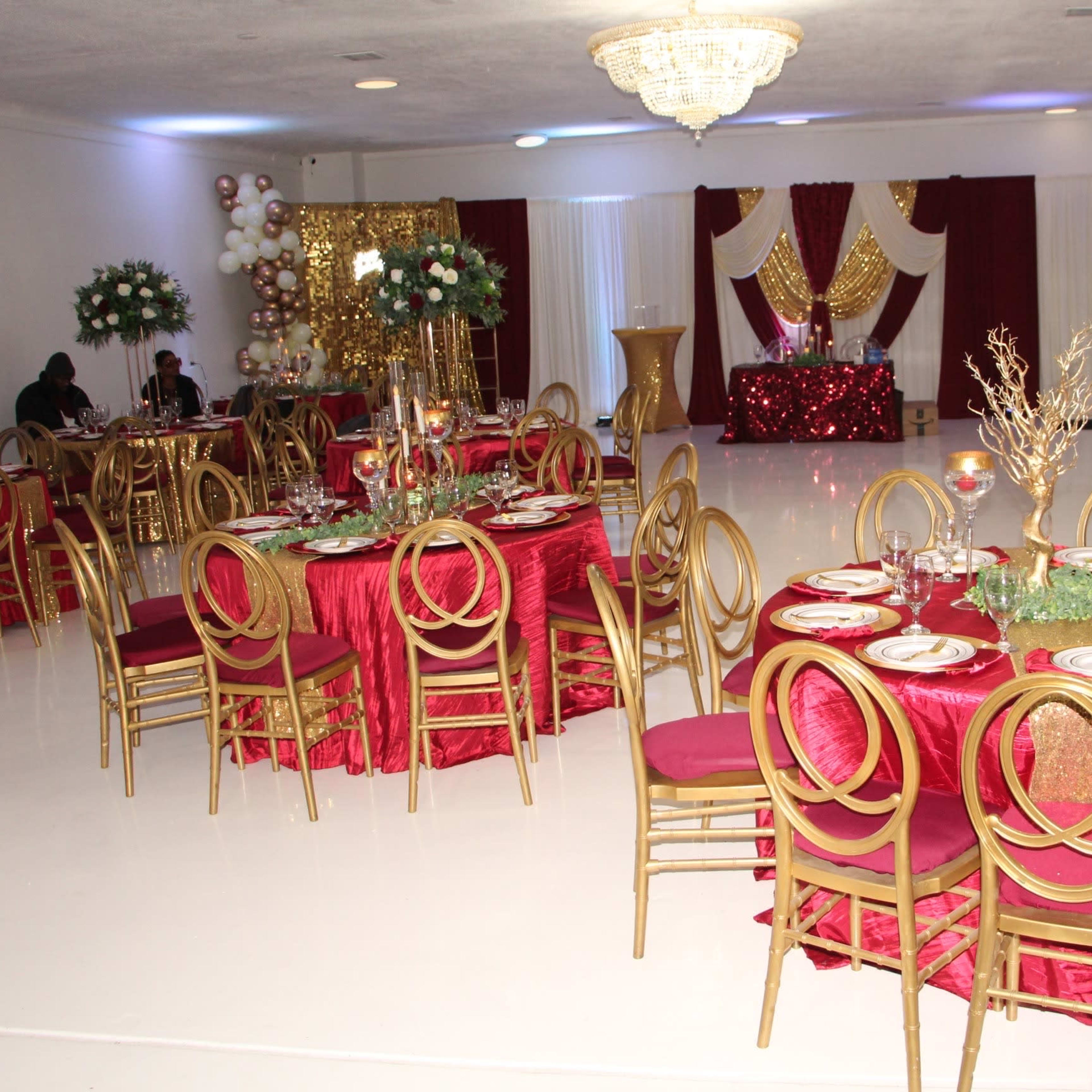 An elegantly decorated event space set for a formal gathering, featuring round tables with red tablecloths and gold chairs, adorned with floral centerpieces and glassware.