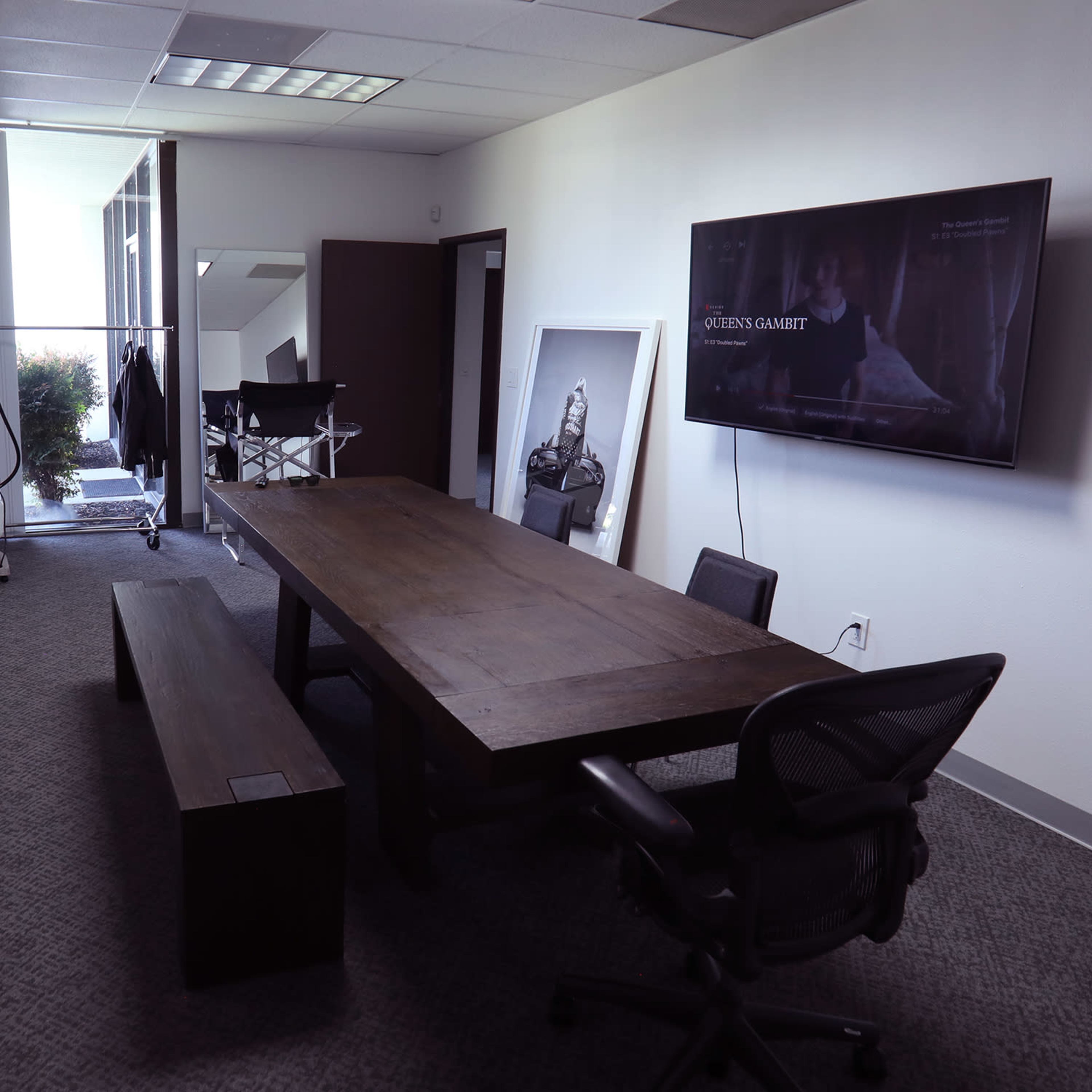 A modern conference room with a large wooden table, black chairs, a television mounted on the wall, and natural light filtering through a window.