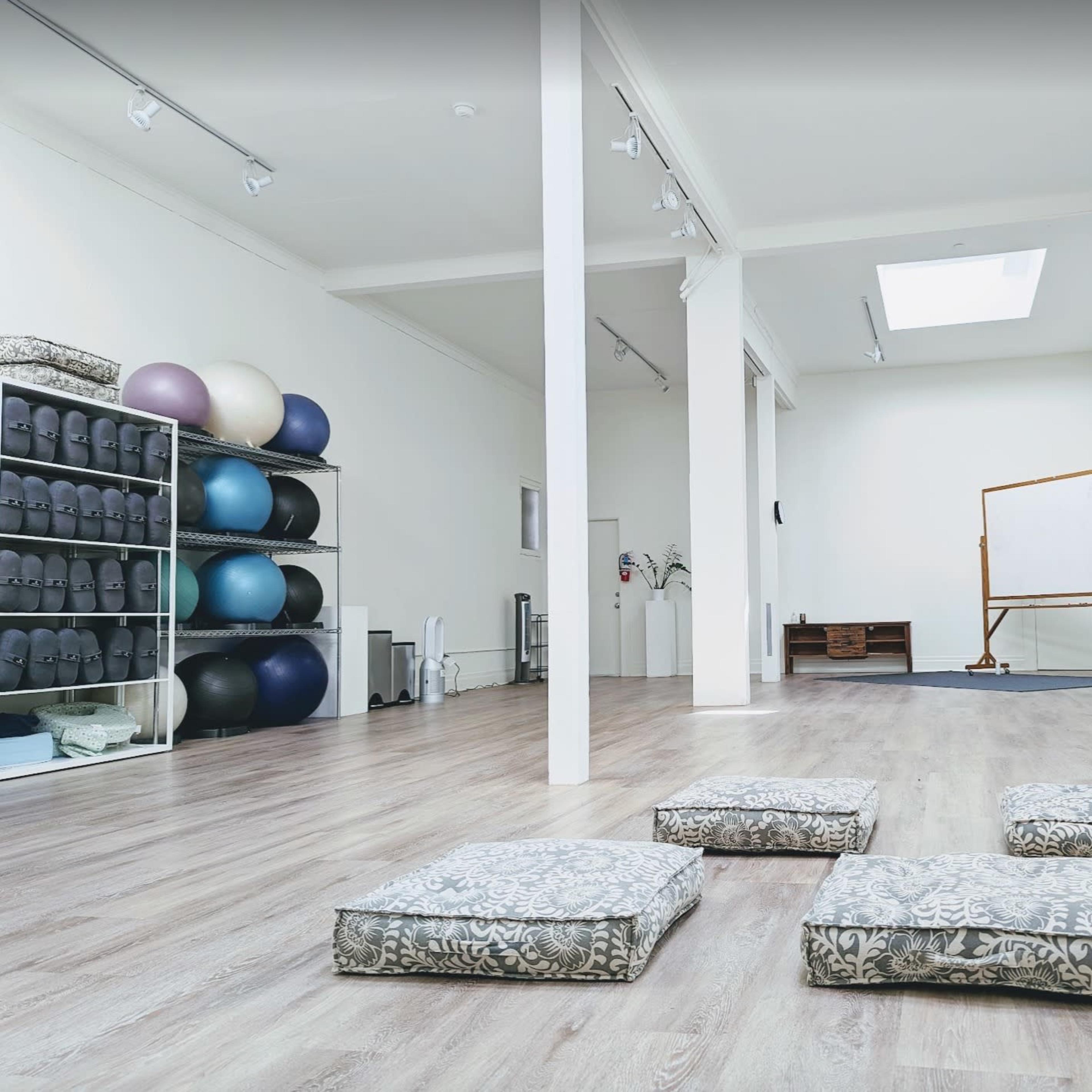A spacious, well-lit room with exercise equipment on shelves, several cushions arranged on the floor, and a whiteboard set up against a wall.