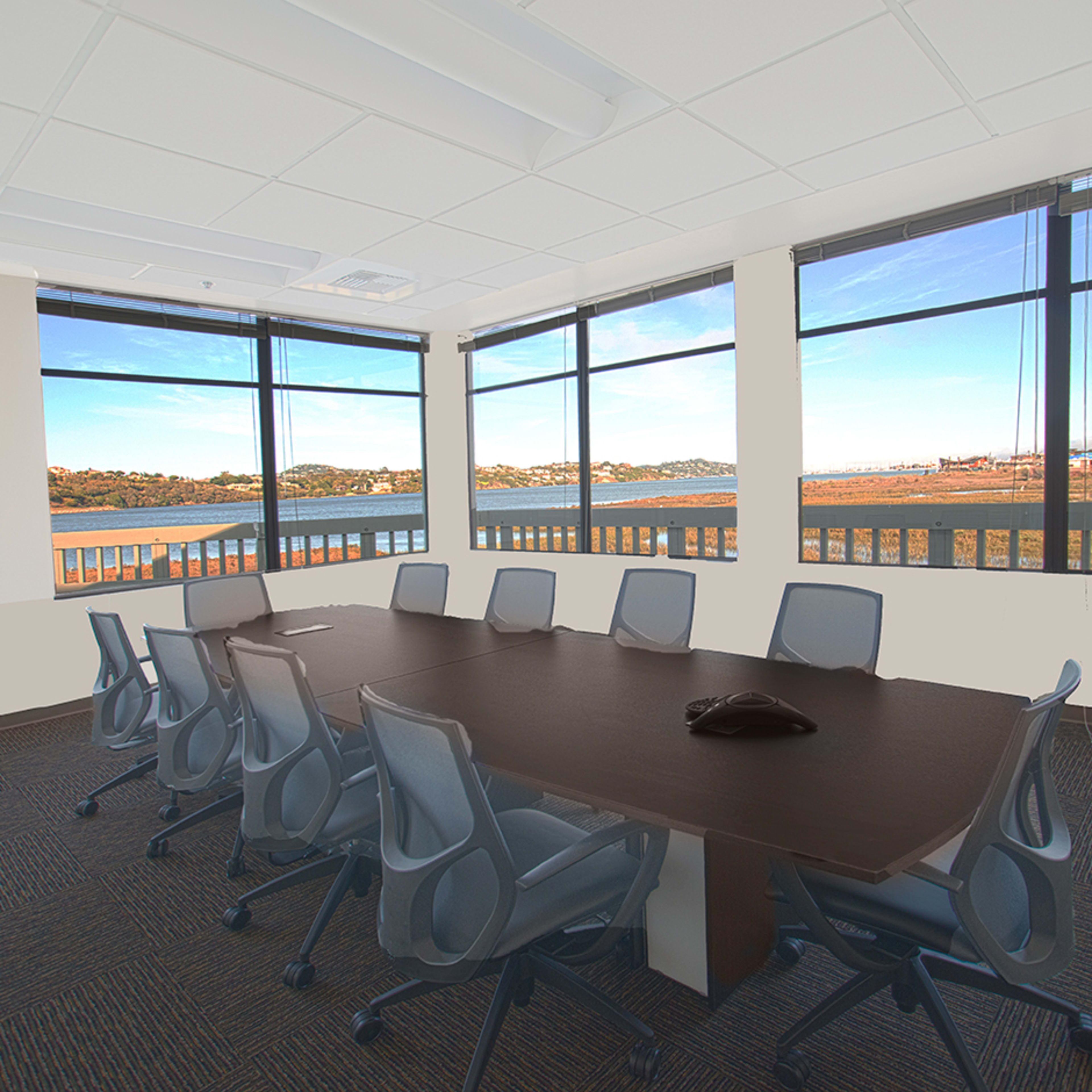 A conference room features a large table surrounded by chairs, with multiple windows offering a view of a landscape by the water.