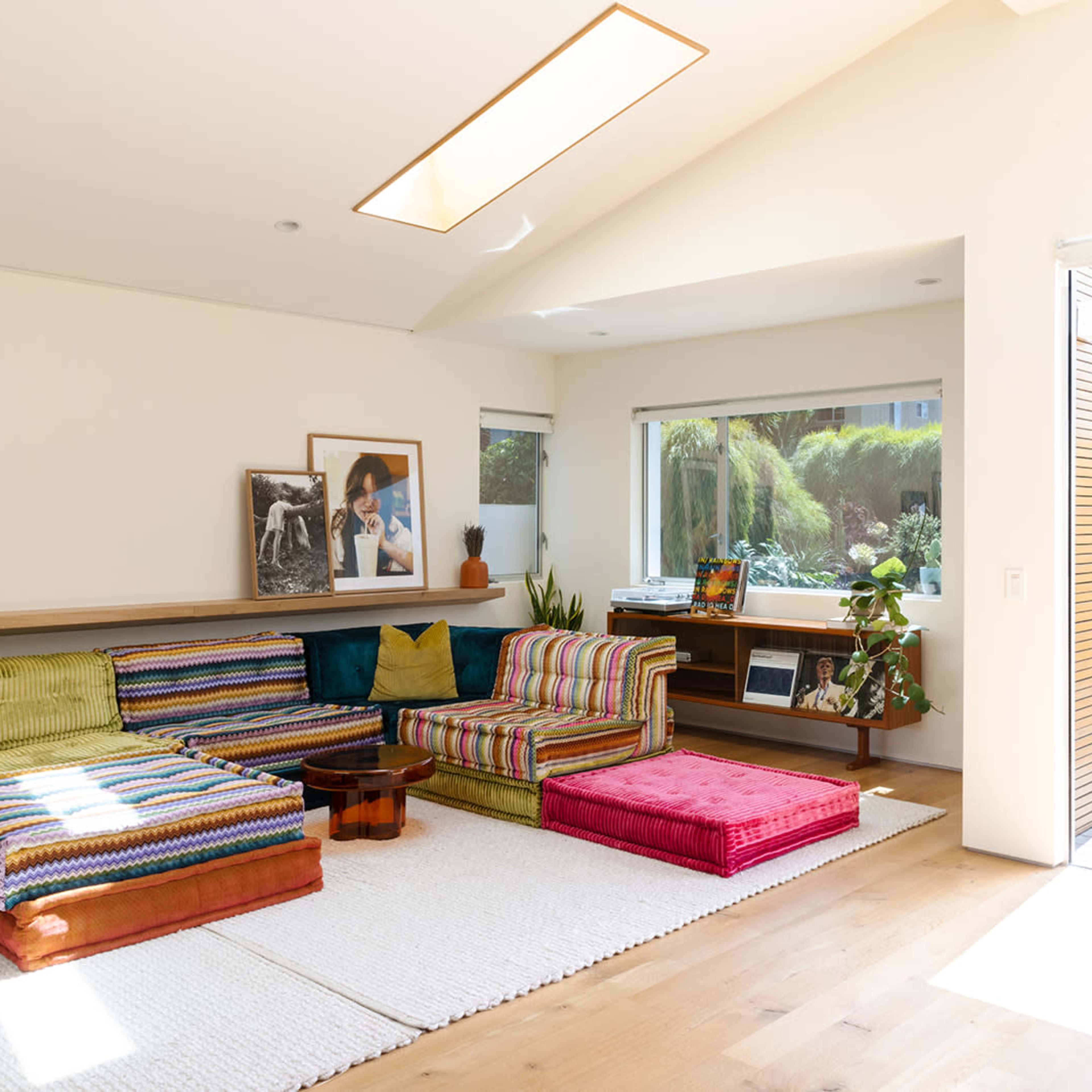 The living room features colorful striped seating, a wooden coffee table, and large windows that open to a green outdoor area.