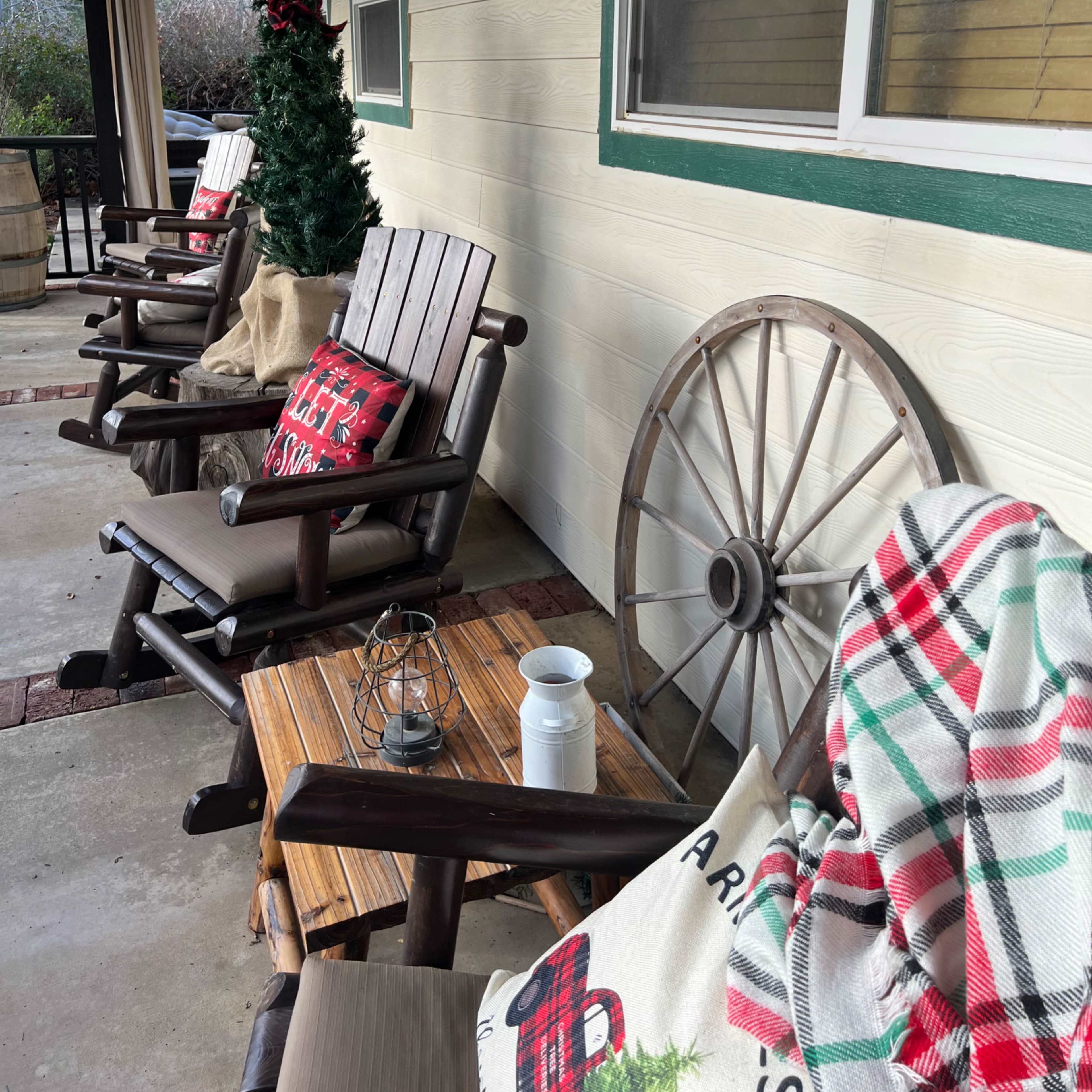 A porch with three wooden rocking chairs, each with a cushion, a coffee table, and holiday decorations, including a small Christmas tree and a wagon wheel.